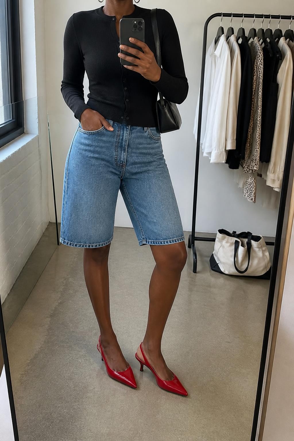 mirror selfie of a Black woman wearing washed medium-blue denim jorts with a relaxed knee-length fit, a fitted black fine-knit cardigan buttoned as a top, red pointed slingback heels with a low heel, a black rectangular shoulder bag and medium silver hoop earrings, wearing exactly the listed outfit with no additional garments or accessories, holding a phone in front of a mirror, cropped at NECK so the face does not show, relaxed confident selfie pose, in a fashion studio mirror corner, natural window light, clothing rack with garments behind and a tote bag resting on the floor, believable casual latest iPhone photo quality.