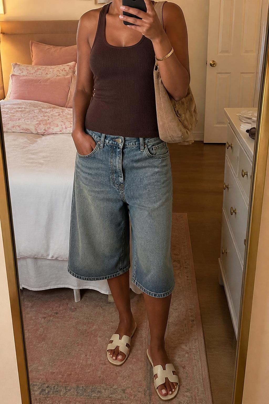 mirror selfie of a Black woman wearing faded blue long denim jorts with a relaxed below-the-knee fit, a fitted chocolate brown ribbed cotton tank, cream flat leather sandals, a tan suede shoulder bag and a simple gold cuff bracelet, wearing exactly the listed outfit with no additional garments or accessories, holding a phone in front of a mirror, cropped at NECK so the face does not show, relaxed casual selfie pose, in a feminine-themed bedroom, evening golden hour, sunglasses placed on a dresser and soft pillows on an unmade bed, natural latest iPhone photo quality.