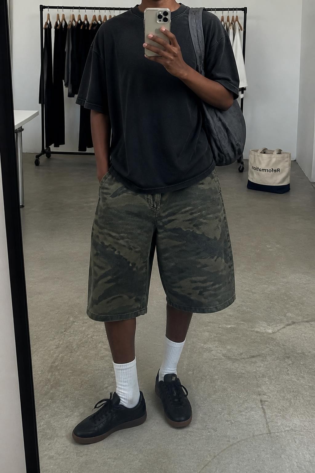 mirror selfie of a Black woman wearing loose green camouflage denim jorts with a knee-length cut, an oversized washed black heavyweight cotton T-shirt with no graphics, white ribbed crew socks, black retro sneakers and a charcoal nylon shoulder bag, wearing exactly the listed outfit with no extra clothing pieces or accessories, holding a phone in front of a mirror, cropped at NECK so the face does not show, relaxed cool-girl selfie pose, in a fashion studio mirror corner, soft indoor lighting, clothing rack with garments behind and a tote bag resting on the floor, natural unstaged latest iPhone photo quality.