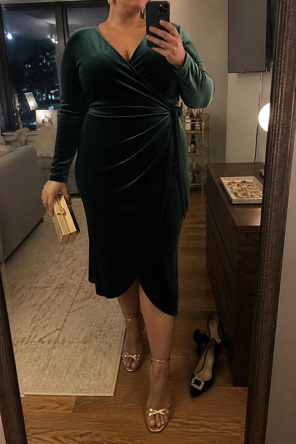 Mirror selfie of a plus size woman wearing an emerald green velvet midi wrap dress with long sleeves and a softly defined waist, metallic gold strappy heels, gold drop earrings, and a small structured gold clutch, clearly showing the exact clothing items, colors, fabrics, and accessories listed, holding a phone in front of a mirror, cropped at the neck so the face does not show, relaxed confident selfie pose, in a stylish city apartment with warm moody lighting, a pair of heels on the floor nearby and a jewelry tray on the dresser, natural unstaged Christmas party appearance, latest iPhone photo quality.