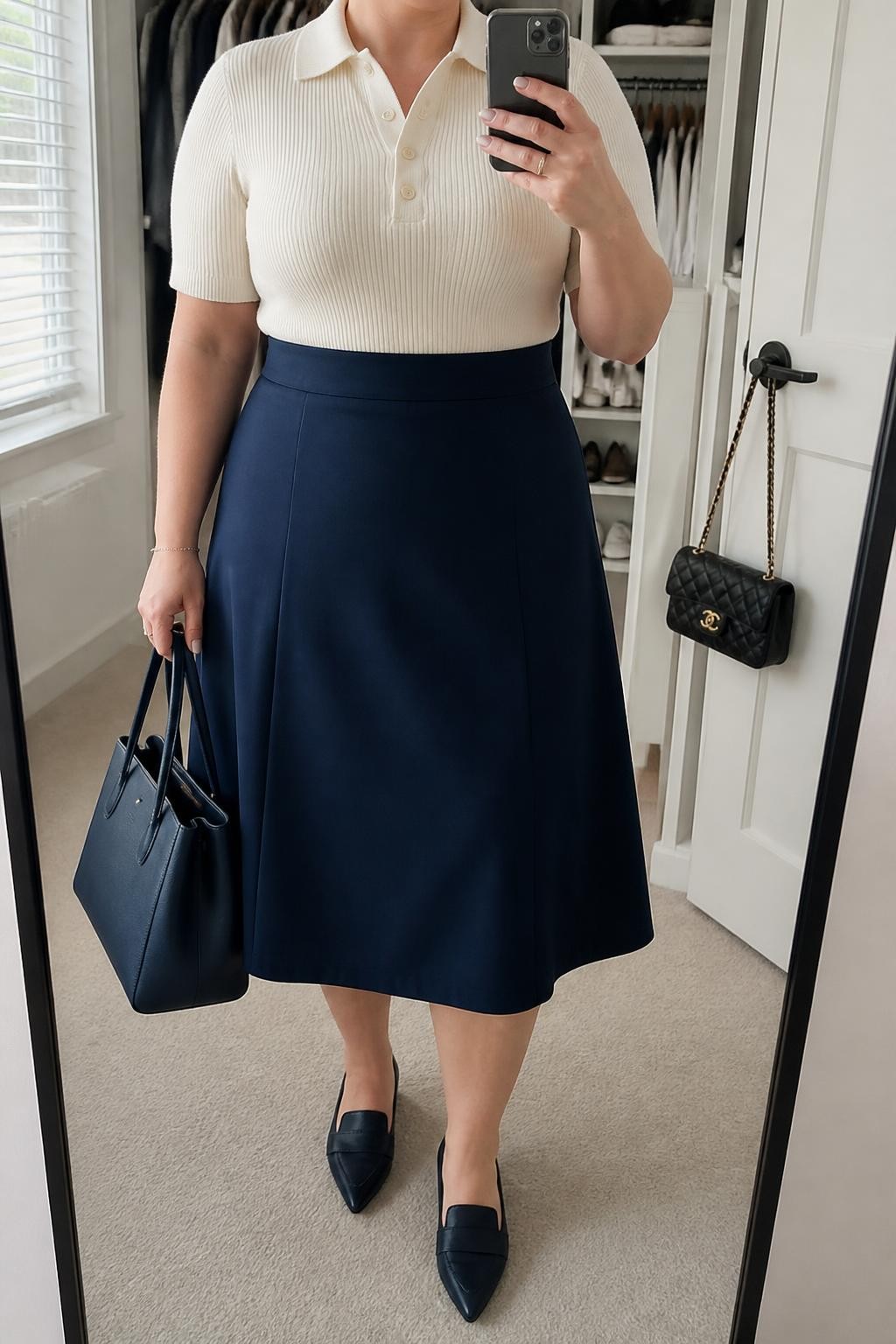 Mirror selfie of a plus size woman wearing a navy high-waisted A-line midi skirt in structured fabric, an ivory fitted short-sleeve knit polo, navy pointed loafers, a structured navy leather tote, and small pearl stud earrings, clearly showing the exact clothing items, navy skirt, ivory knit, colors, footwear, and accessories listed, holding a phone in front of a mirror, cropped at the neck so her face does not appear, relaxed neat selfie pose, in a walk-in closet mirror with natural window light, an open closet with clothes visible and a handbag hanging on a door handle, realistic business-casual interview outfit, latest iPhone photo quality.