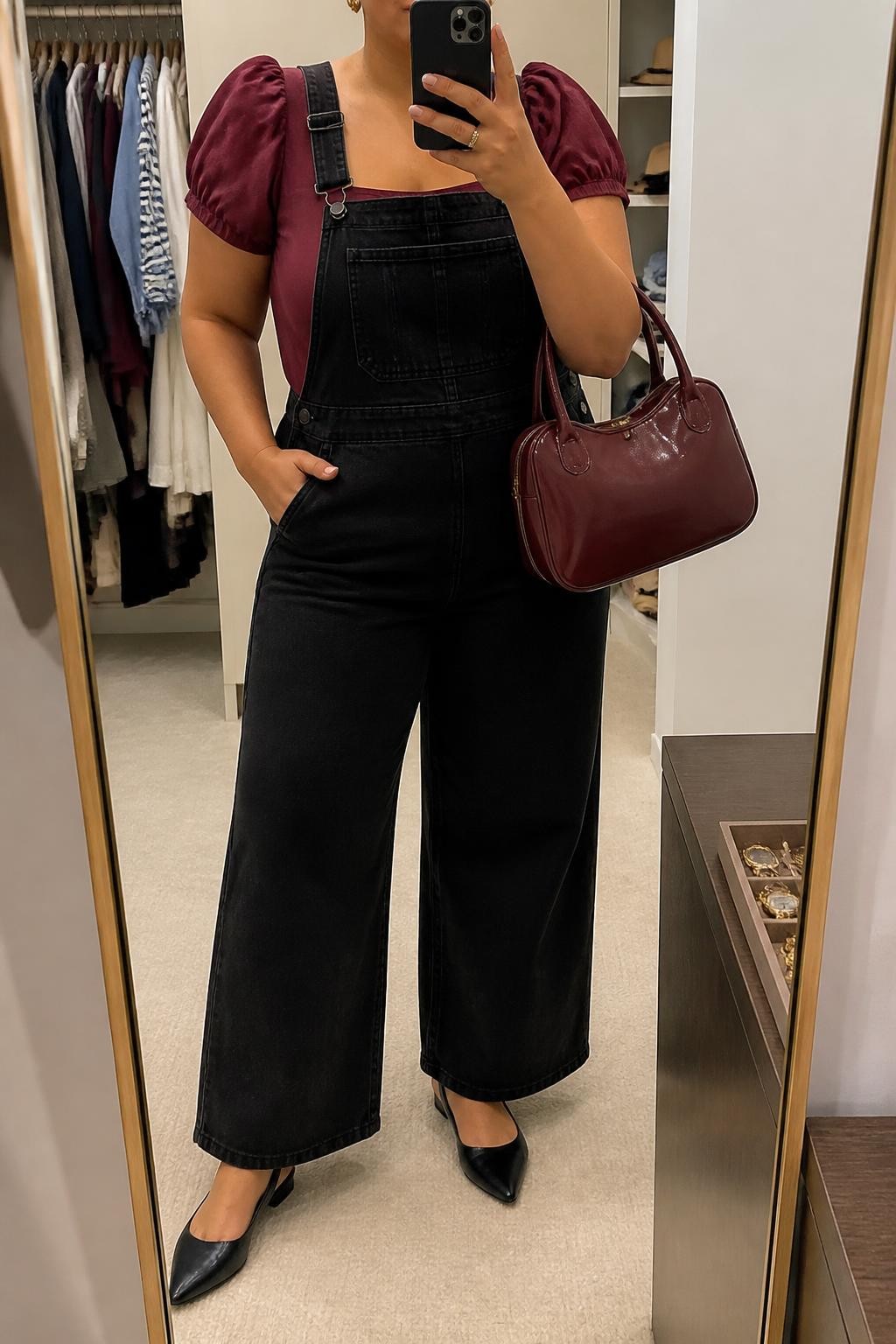 Mirror selfie of a plus size woman wearing black wide-leg denim overalls with adjustable straps and a square bib, layered over a burgundy cotton blouse with short puff sleeves and a square neckline, black pointed slingback flats, a burgundy patent handbag, and small gold stud earrings, clearly showing the exact clothing items, black denim, burgundy blouse, sleeve shape, footwear, and accessories listed, holding a phone in front of a mirror, cropped at the neck so the face is hidden, relaxed feminine selfie pose, in a walk-in closet mirror with soft indoor lighting, an open closet with clothes visible and a jewelry tray on the vanity, realistic brunch outfit, latest iPhone photo quality.