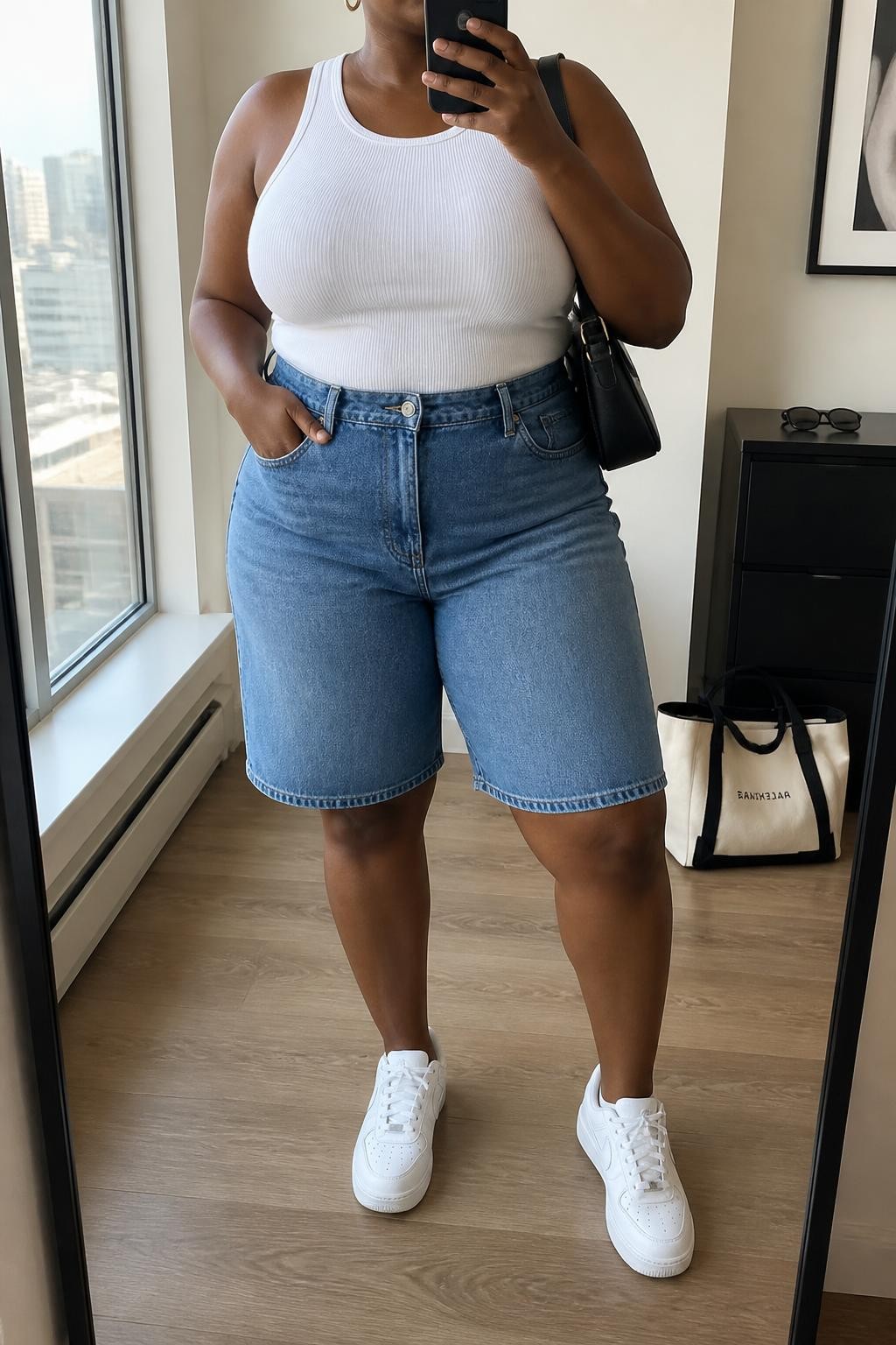 mirror selfie of a plus size Black woman wearing high-waisted medium-wash blue denim jorts with a relaxed knee-length fit, a fitted white ribbed cotton tank top, white retro sneakers, a black leather shoulder bag and medium gold hoop earrings, wearing exactly the listed clothing and accessories with no extra garments, holding a phone in front of a mirror, cropped at NECK so the face does not show, relaxed confident selfie pose, in a stylish city apartment, natural window light, sunglasses placed on a dresser and a tote bag resting on the floor, realistic body proportions and natural denim folds, latest iPhone photo quality.