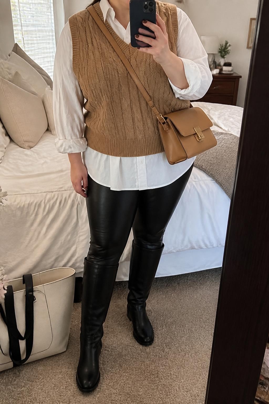 Mirror selfie of a plus size woman wearing a camel cable-knit sweater vest layered over an oversized white cotton button-down shirt, black high-waisted faux-leather leggings, black knee-high riding boots, and a structured camel crossbody bag, clearly showing the exact clothing layers, camel knit, white shirt, black leggings, footwear, and accessories listed, holding a phone in front of a mirror, cropped at the neck so her face does not appear, relaxed preppy selfie pose, in a cozy bedroom mirror setup with natural window light, soft pillows on an unmade bed and a tote bag resting on the floor, realistic relaxed Thanksgiving outfit, latest iPhone photo quality.
