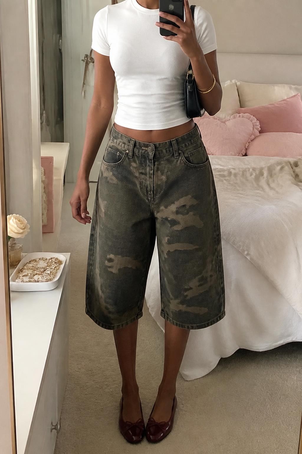 mirror selfie of a Black woman wearing oversized woodland camo denim jorts with a relaxed below-the-knee fit, a fitted white cotton baby tee, burgundy leather ballet flats, a black mini shoulder bag and small gold hoop earrings, wearing exactly the listed outfit with no added clothing pieces, holding a phone in front of a mirror, cropped at NECK so the face does not show, relaxed casual selfie pose, in a feminine-themed bedroom, soft indoor lighting, soft pillows on an unmade bed and a jewelry tray on the vanity, believable everyday styling, latest iPhone photo quality.