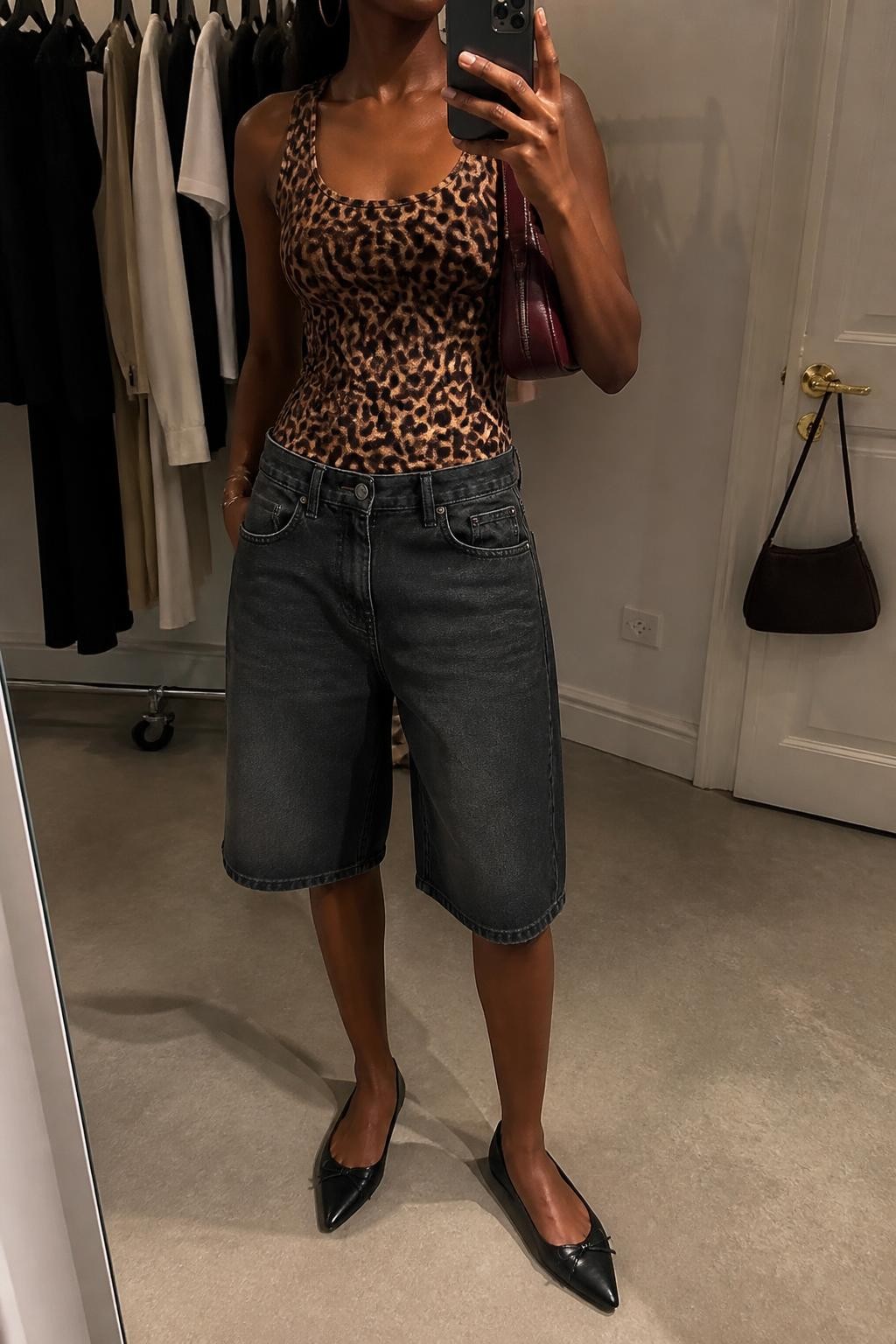 mirror selfie of a Black woman wearing washed black baggy knee-length denim jorts, a fitted leopard print tank top in warm brown and black tones, pointed black leather flats, a small burgundy leather shoulder bag and medium gold hoop earrings, wearing exactly the listed clothing and accessories without added garments, holding a phone in front of a mirror, cropped at NECK so the face does not show, relaxed cool-girl selfie pose, in a fashion studio mirror corner, warm moody lighting, clothing rack with garments behind and a handbag hanging on a door handle, natural unstaged styling and latest iPhone photo quality.