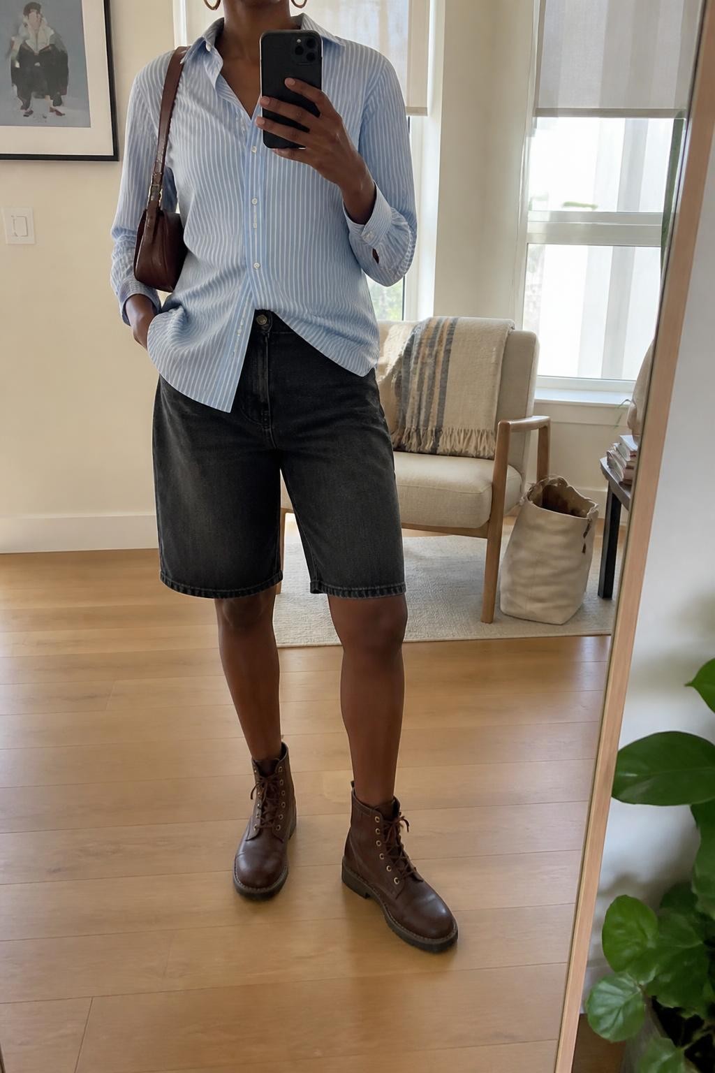 mirror selfie of a Black woman wearing straight black Bermuda denim jorts, a fitted pale blue and white striped cotton button-up shirt loosely tucked at the front, dark brown leather lace-up boots, a brown leather shoulder bag and small gold hoop earrings, wearing exactly the listed outfit with no added clothing pieces, holding a phone in front of a mirror, cropped at NECK so the face does not show, relaxed casual selfie pose, in a stylish city apartment, natural window light, tote bag resting on the floor and a folded blanket on a chair, believable everyday styling, latest iPhone photo quality.