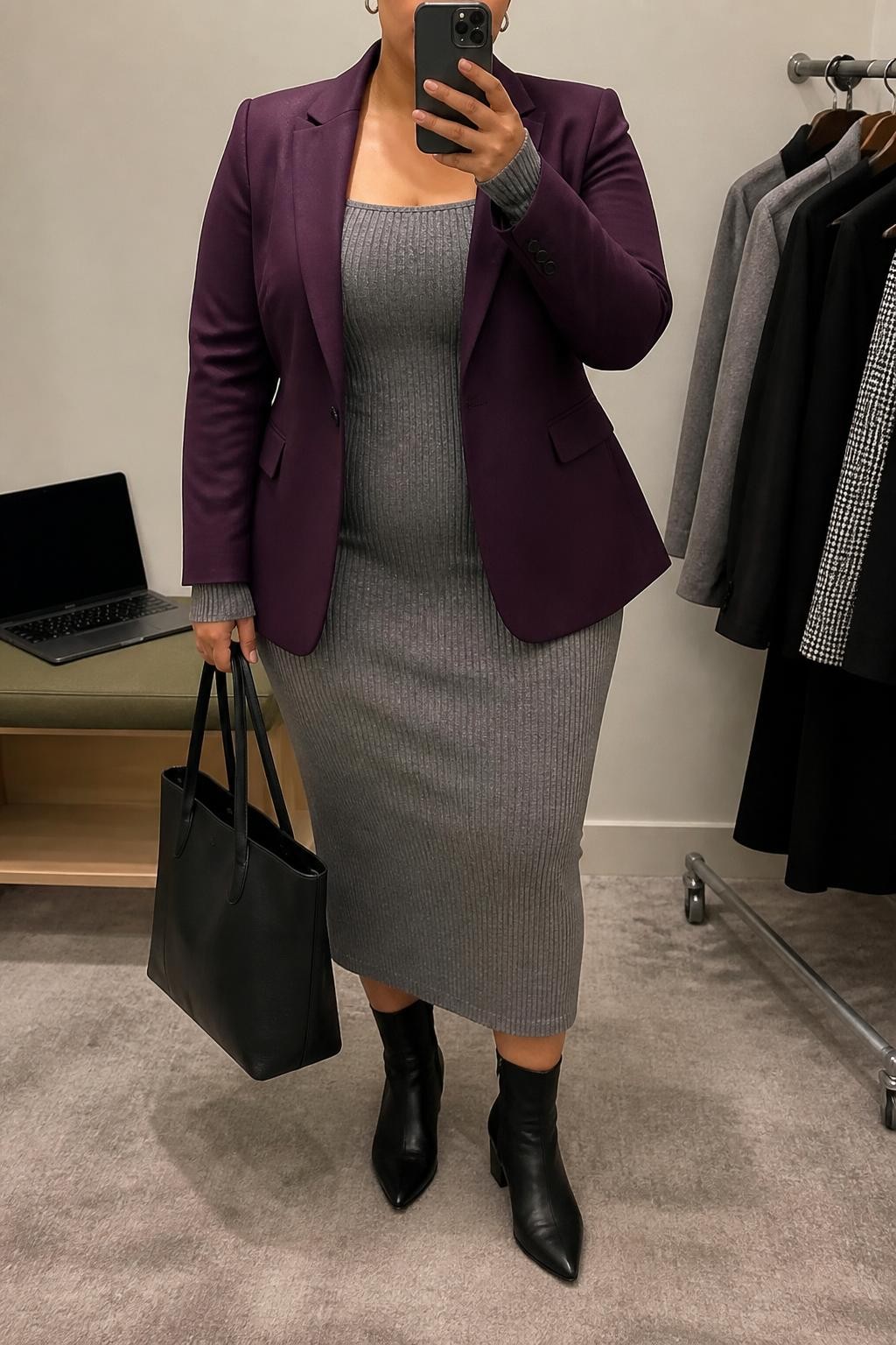 Mirror selfie of a plus size woman wearing a plum purple tailored blazer over a fitted grey knit midi dress with long sleeves and a modest square neckline, black pointed ankle boots with low block heels, a structured black leather tote, and small silver hoop earrings, clearly showing the exact clothing items, plum blazer, grey dress, colors, footwear, and accessories listed, holding a phone in front of a mirror, cropped at the neck so the face is hidden, relaxed professional selfie pose, in a minimal dressing room with soft indoor lighting, a laptop on a bench and a clothing rack with garments behind her, realistic polished interview appearance, latest iPhone photo quality.
