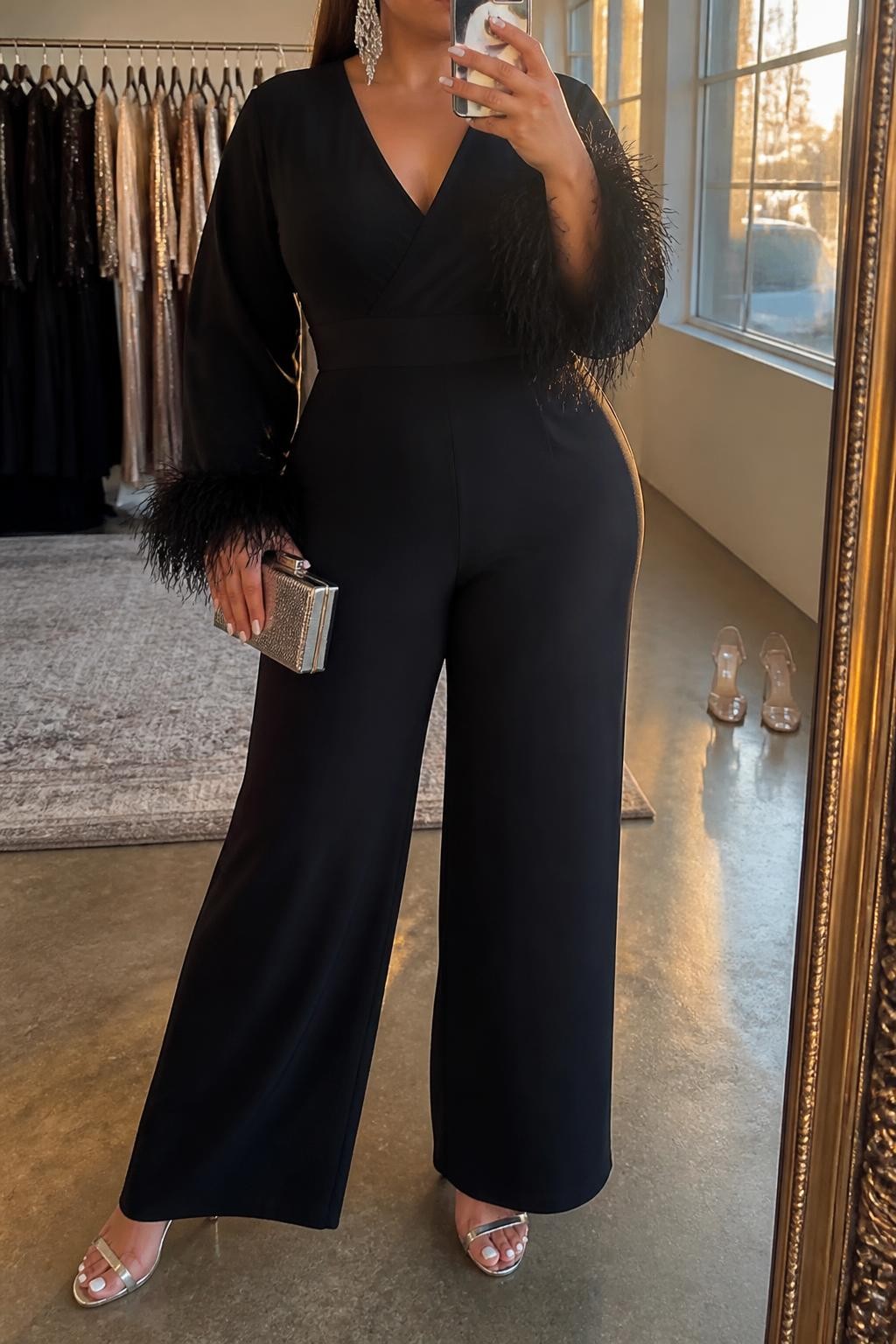 Mirror selfie of a plus size woman wearing a black wide-leg jumpsuit with a defined waist and delicate feather trim around the sleeve cuffs, silver crystal chandelier earrings, silver metallic heels, and a small silver box clutch, clearly showing the exact outfit pieces, colors, feather details, fabrics, and accessories listed, holding a phone in front of a mirror, cropped at the neck so her face is hidden, confident relaxed selfie stance, in a fashion studio mirror corner with evening golden-hour lighting, a clothing rack with garments behind her and a pair of heels on the floor nearby, natural glamorous party setting, latest iPhone photo quality.