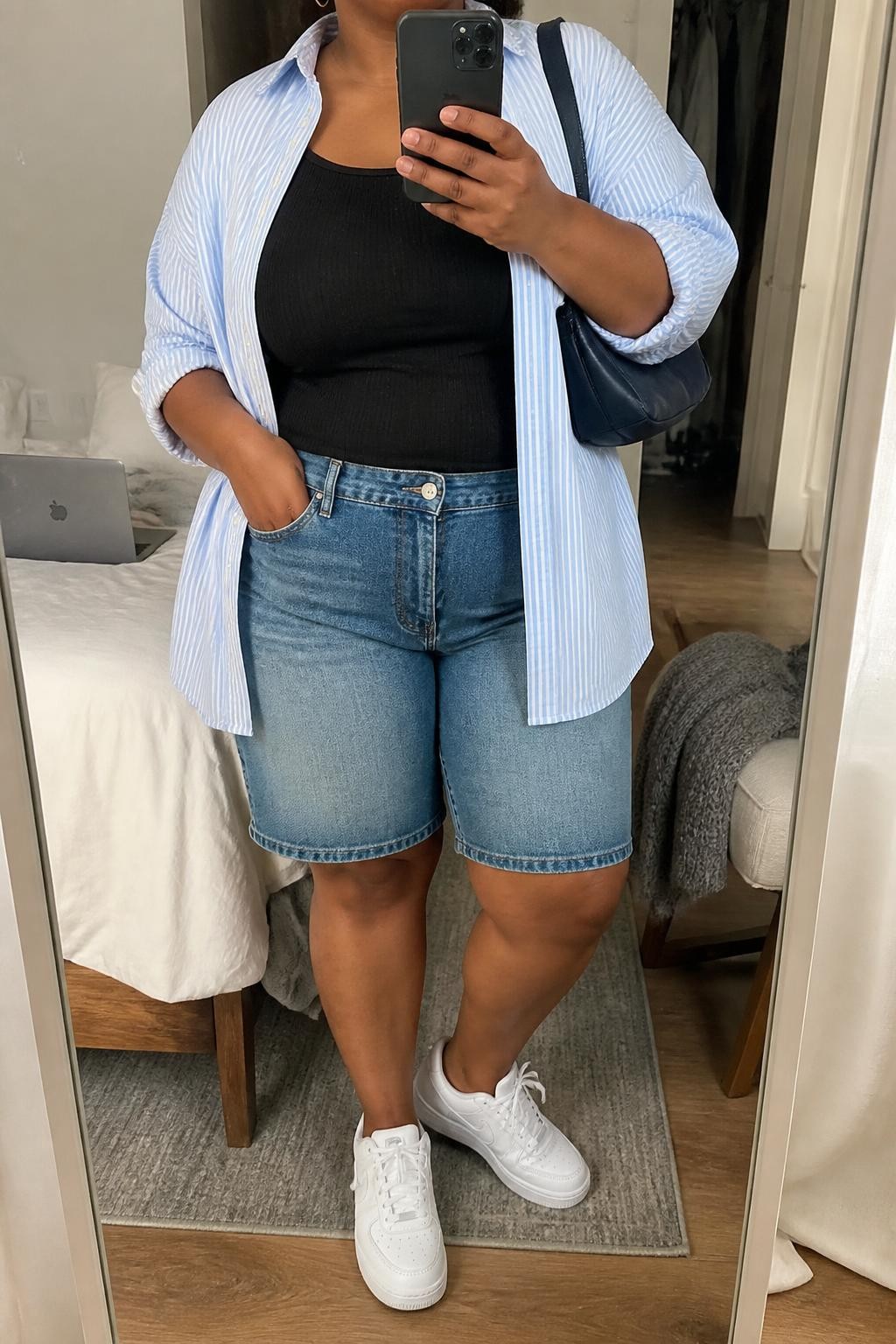 mirror selfie of a plus size Black woman wearing medium-wash straight-cut denim jorts, an oversized pale blue and white striped cotton shirt worn open over a fitted black ribbed tank, white leather sneakers, a navy shoulder bag and small silver hoop earrings, wearing exactly the listed outfit with no added garments, holding a phone in front of a mirror, cropped at NECK so the face does not show, relaxed casual selfie pose, in a cozy bedroom mirror setup, natural window light, laptop on the bed and a folded blanket on a chair, believable everyday styling, latest iPhone photo quality.