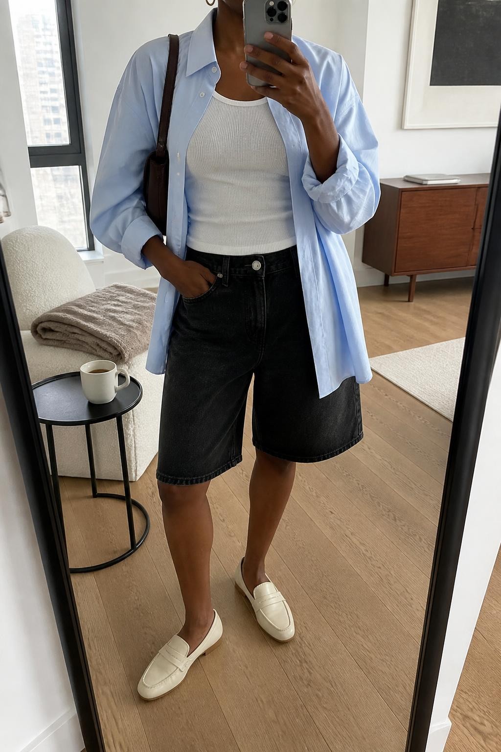 mirror selfie of a Black woman wearing washed black relaxed denim jorts with a knee-length cut, a pale blue oversized cotton button-up shirt worn open over a fitted white ribbed tank, cream leather loafers, a dark brown shoulder bag and small gold hoop earrings, wearing exactly the listed clothing and accessories with no extra pieces, holding a phone in front of a mirror, cropped at NECK so the face does not show, relaxed casual selfie pose, in a stylish city apartment, natural window light, coffee mug on a side table and a folded blanket on a chair, believable latest iPhone photo quality.