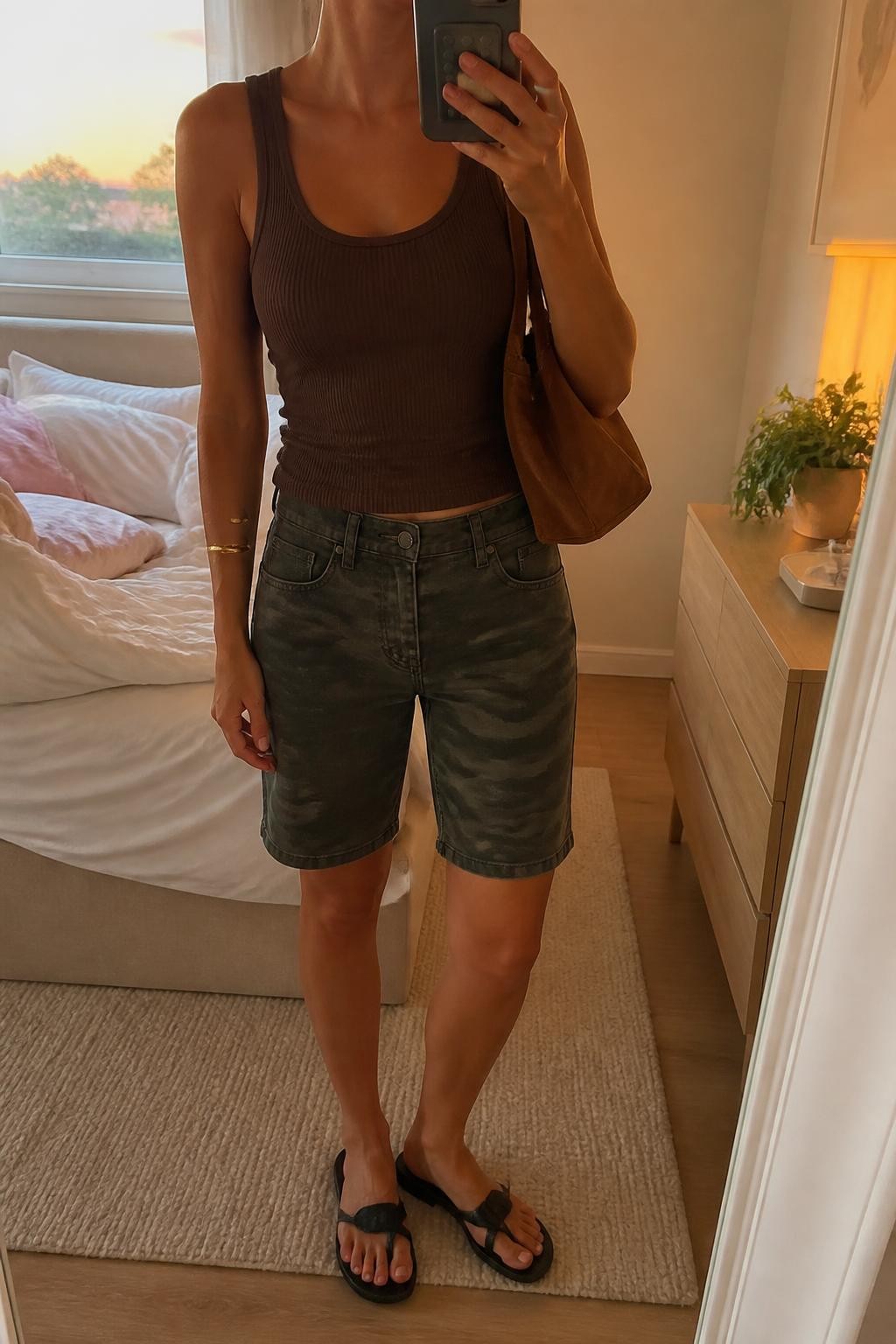 mirror selfie of a woman with light olive skin wearing dark olive camouflage denim jorts with a relaxed straight cut, a fitted chocolate brown ribbed tank top, black flat leather sandals, a brown suede shoulder bag and a simple gold cuff bracelet, wearing exactly the outfit listed without extra clothing pieces, holding a phone in front of a mirror, cropped at NECK so the face does not show, relaxed casual selfie pose, in a feminine-themed bedroom, evening golden hour, soft pillows on an unmade bed and a small plant on a dresser, natural casual latest iPhone photo quality.