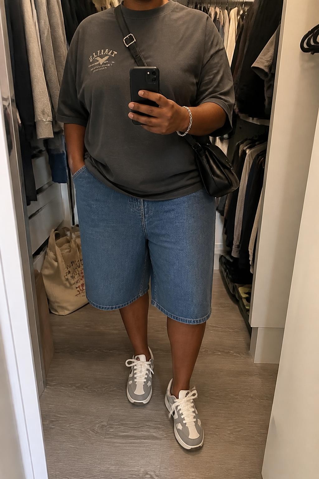 mirror selfie of a plus size Black woman wearing baggy blue denim jorts with a loose knee-length cut, an oversized charcoal gray cotton graphic T-shirt with a small faded cream graphic, gray and white retro sneakers, a black nylon crossbody bag and a silver chain bracelet, wearing exactly the listed outfit with no additional clothing or accessories, holding a phone in front of a mirror, cropped at NECK so the face does not show, relaxed street-style selfie pose, in a walk-in closet mirror, soft indoor lighting, open closet with clothes visible and a tote bag resting on the floor, natural slightly relaxed styling, latest iPhone photo quality.