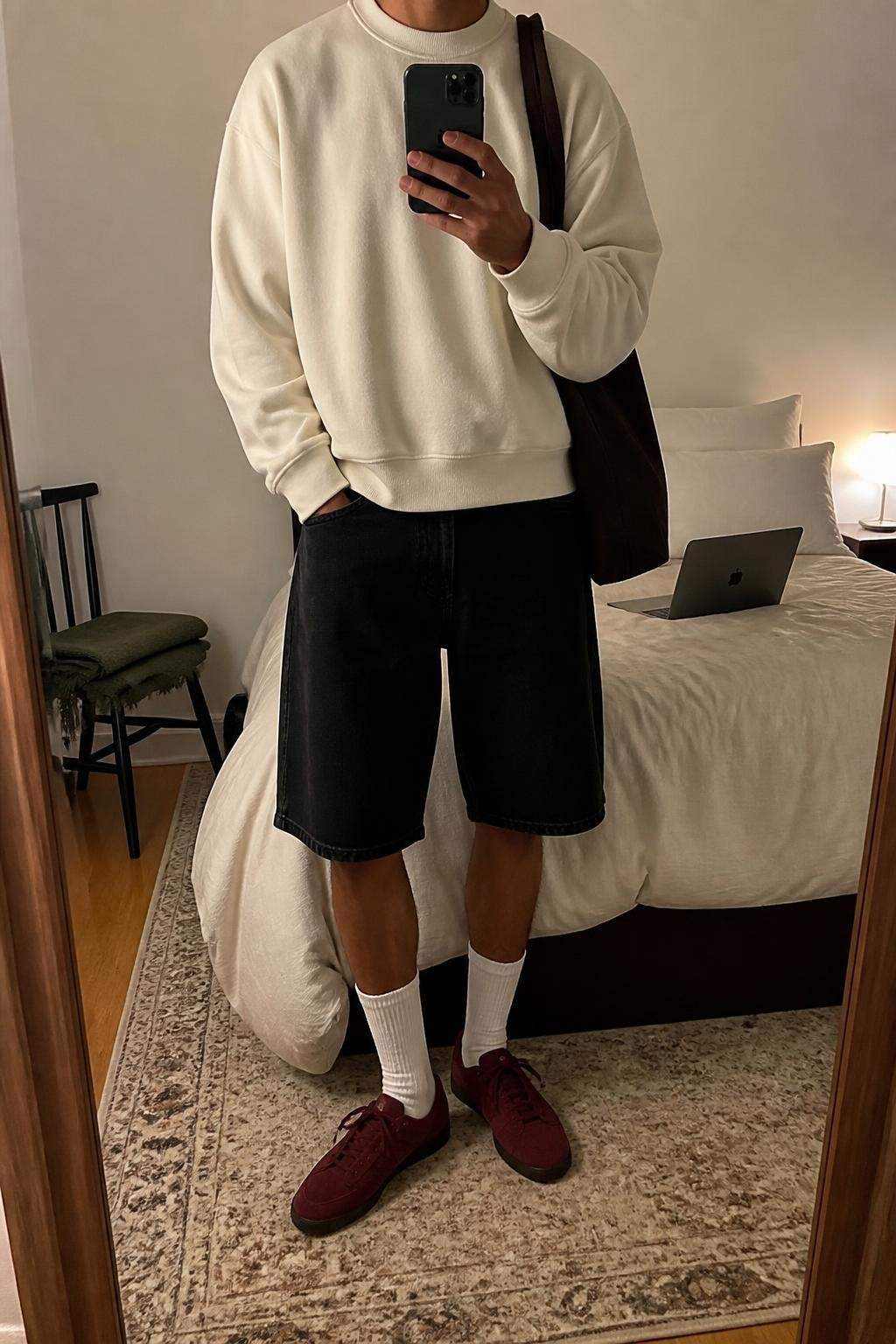 mirror selfie of a man with warm tan skin wearing clean knee-length black denim jorts, an oversized cream cotton sweatshirt with no graphics, white ribbed crew socks, burgundy low-profile sneakers and a dark brown canvas tote bag, wearing exactly the outfit listed and no additional pieces, holding a phone in front of a mirror, cropped at NECK so the face does not show, relaxed masculine selfie pose, in a cozy bedroom mirror setup, soft indoor lighting, laptop on the bed and folded blanket on a chair, natural casual appearance, latest iPhone photo quality.