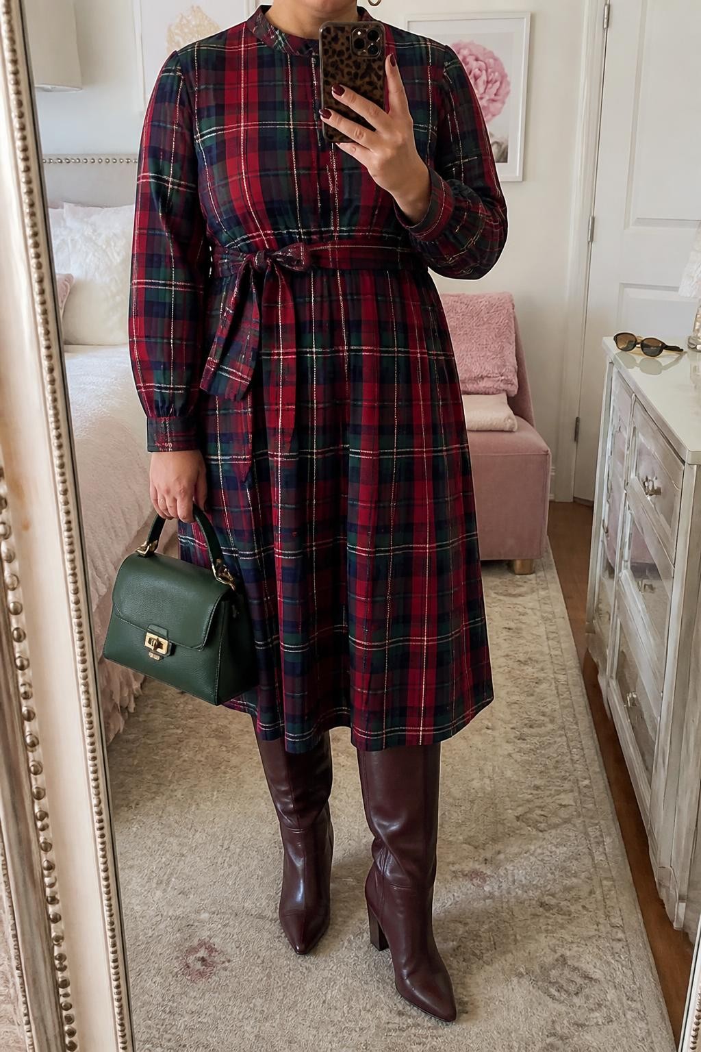 Mirror selfie of a plus size woman wearing a red, green, and navy plaid midi dress with long sleeves and a softly belted waist, burgundy leather knee-high boots, a dark green top-handle bag, and small gold hoop earrings, clearly showing the exact dress pattern, colors, fabrics, accessories, and footwear listed, holding a phone in front of a mirror, cropped at the neck so the face does not appear, relaxed casual selfie pose, in a feminine-themed bedroom with soft indoor lighting, a folded blanket on a chair and sunglasses placed on a dresser, realistic festive daytime outfit, latest iPhone photo quality.