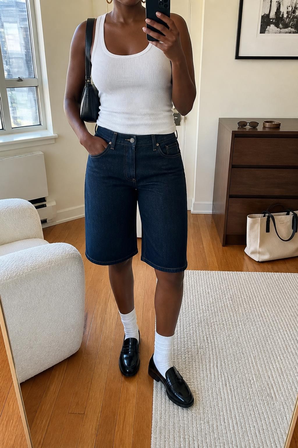 mirror selfie of a Black woman wearing dark blue straight-cut denim jorts ending near the knee, a fitted white ribbed cotton tank top, polished black leather loafers, white ribbed crew socks, a compact black leather shoulder bag and small gold hoop earrings, wearing exactly the listed clothing and accessories with no additional garments, holding a phone in front of a mirror, cropped at NECK so the face does not show, relaxed casual selfie pose, in a stylish city apartment, natural window light, sunglasses placed on a dresser and a tote bag resting on the floor, realistic denim folds and natural proportions, latest iPhone photo quality.