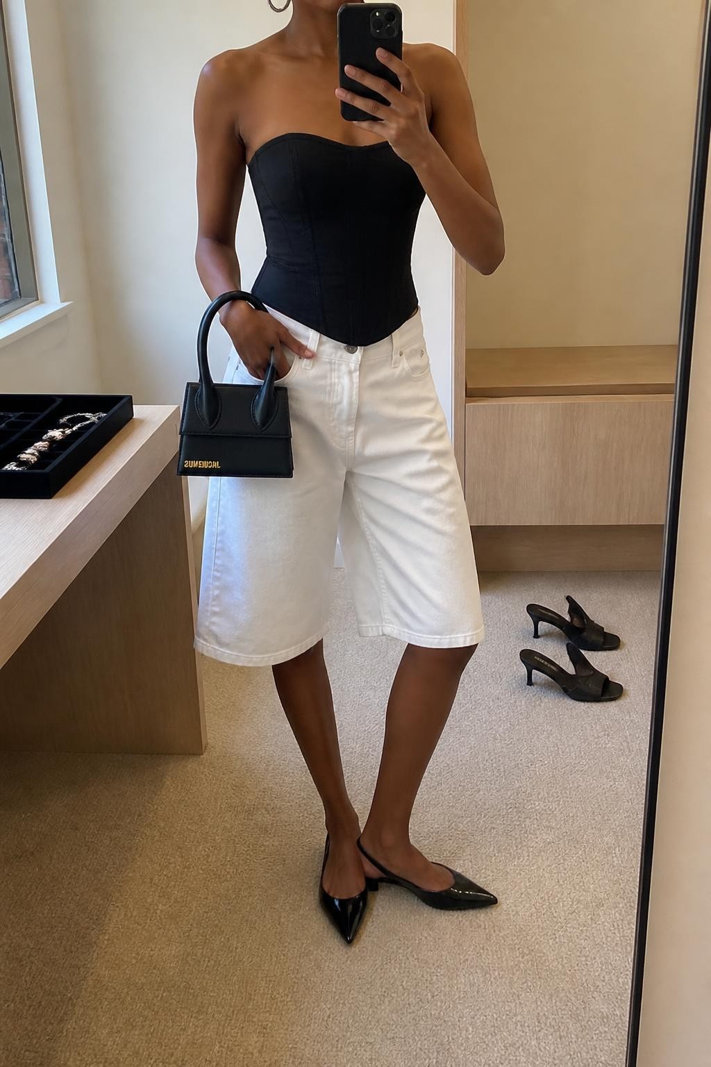 mirror selfie of a Black woman wearing baggy white denim jorts with a loose knee-length fit, a fitted black cotton corset-inspired top with clean seam detailing, black pointed slingback flats, a structured black mini handbag and medium silver hoop earrings, wearing exactly the listed clothing and accessories with no additional garments, holding a phone in front of a mirror, cropped at NECK so the face does not show, relaxed confident selfie pose, in a minimal dressing room, natural window light, pair of heels on the floor nearby and a jewelry tray on the vanity, realistic polished casual styling, latest iPhone photo quality.