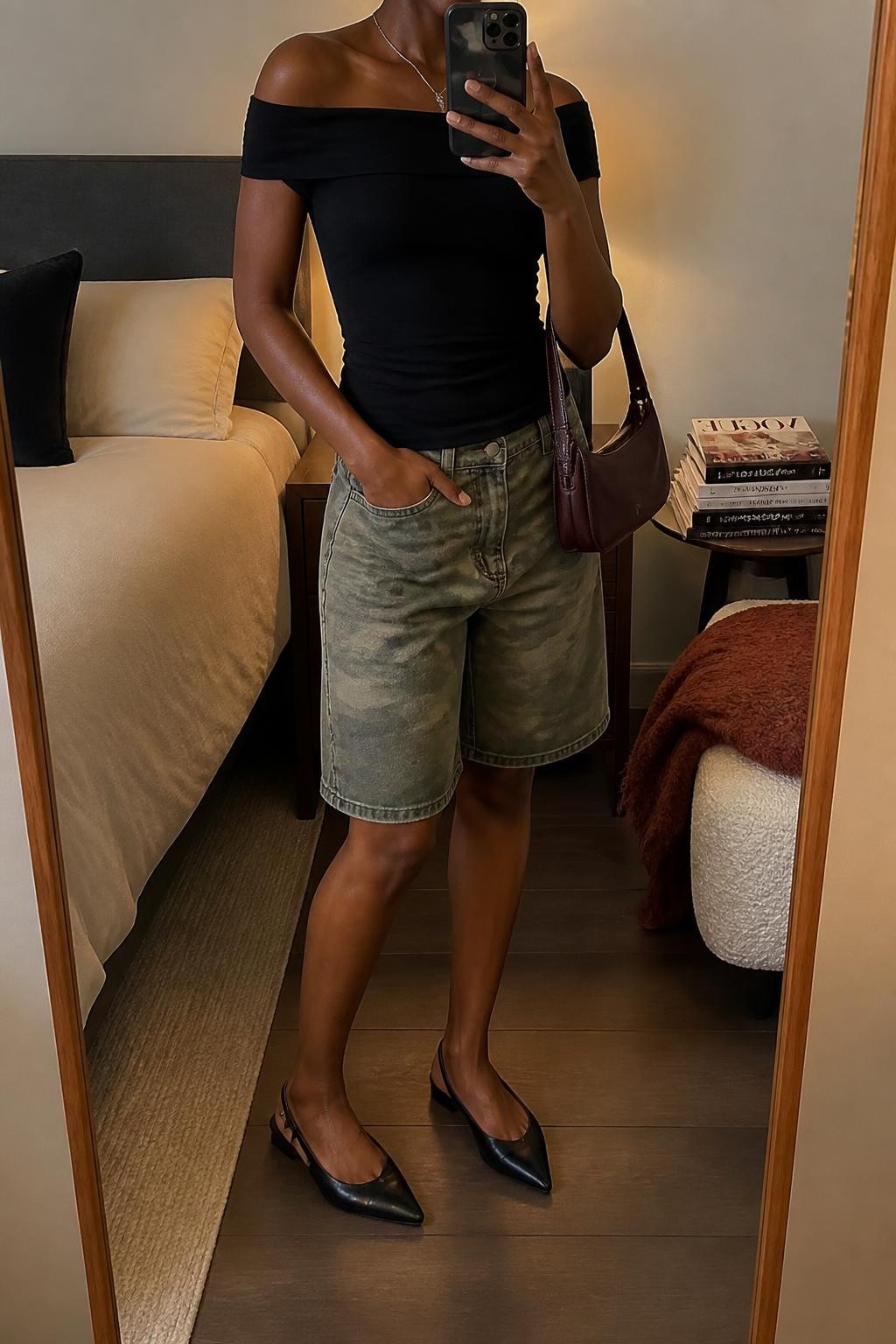 mirror selfie of a Black woman wearing washed green camouflage denim jorts with a loose knee-length cut, a fitted black off-shoulder stretch cotton top, pointed black slingback flats, a burgundy mini shoulder bag and a thin silver pendant necklace, wearing exactly the listed outfit with no extra garments, holding a phone in front of a mirror, cropped at NECK so the face does not show, relaxed casual selfie pose, in a cozy bedroom mirror setup, warm moody lighting, folded blanket on a chair and a stack of fashion magazines on a side table, realistic latest iPhone photo quality.