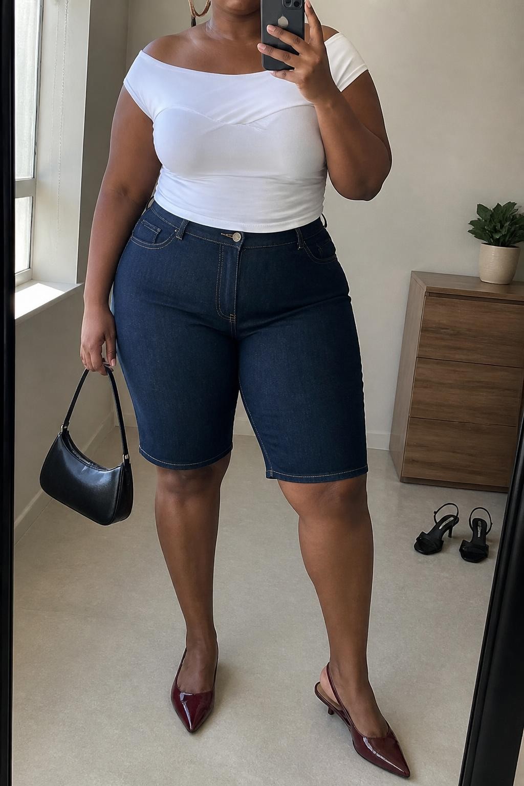 mirror selfie of a plus size Black woman wearing dark blue structured denim jorts with a clean knee-length fit, a fitted white off-shoulder stretch cotton top, burgundy slingback flats, a black mini handbag and medium silver hoop earrings, wearing exactly the listed outfit with no extra garments, holding a phone in front of a mirror, cropped at NECK so the face does not show, relaxed polished selfie pose, in a minimal dressing room, natural window light, a small plant on a dresser and a pair of heels on the floor nearby, believable latest iPhone photo quality.