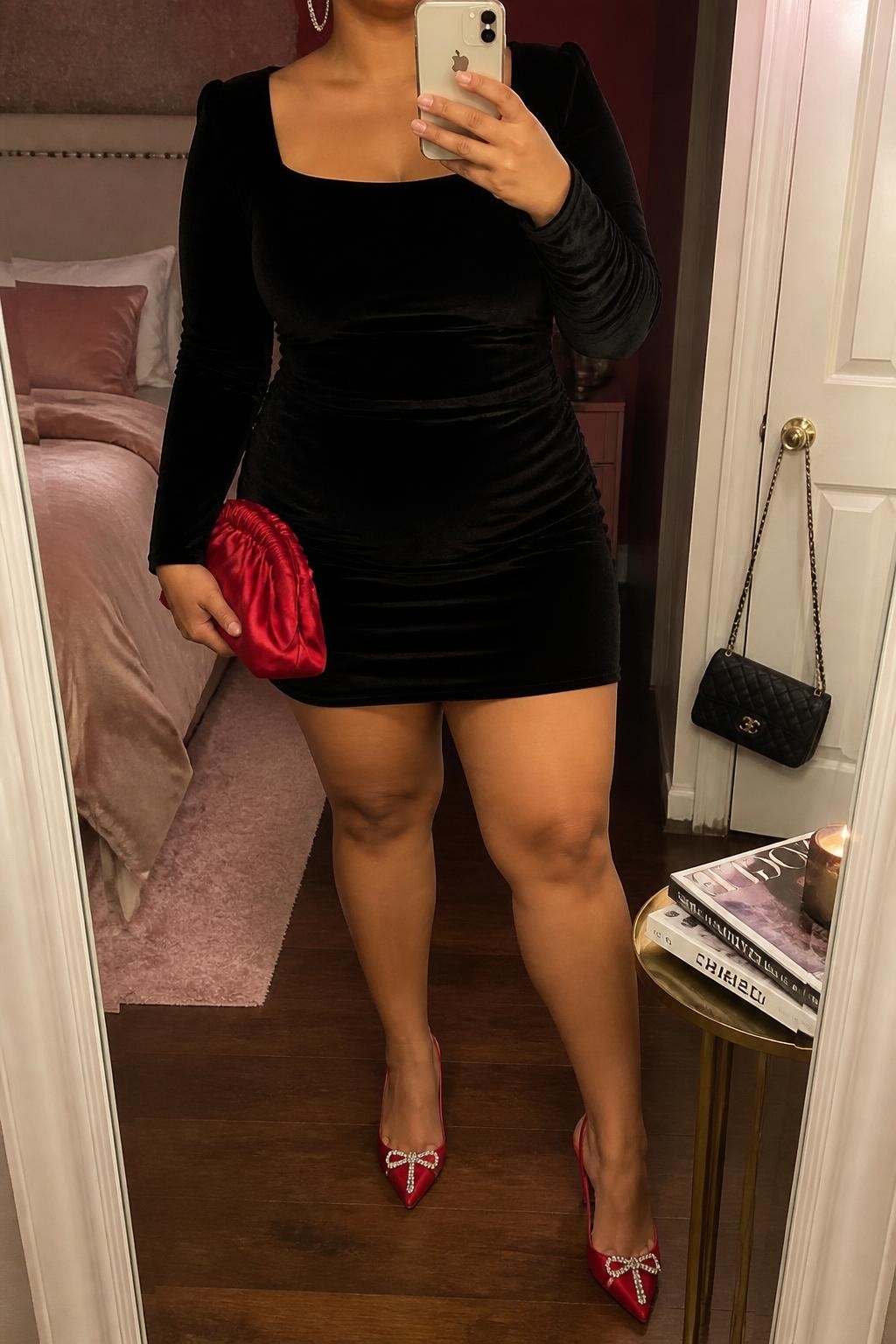 Mirror selfie of a plus size woman wearing a black velvet long-sleeve mini dress with a square neckline and a softly fitted waist, red pointed heels decorated with small crystal details, a red satin clutch, and thin crystal hoop earrings, clearly showing the exact clothing items, colors, velvet texture, crystal details, and accessories listed, holding a phone in front of a mirror, cropped at the neck so the face does not appear, relaxed confident selfie pose, in a feminine-themed bedroom with warm moody lighting, a stack of fashion magazines on a side table and a handbag hanging on a door handle, realistic festive party scene, latest iPhone photo quality.