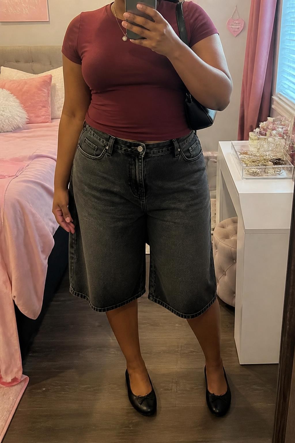 mirror selfie of a plus size Black woman wearing baggy washed black denim jorts with a loose knee-length cut, a fitted burgundy cotton baby tee, black leather ballet flats, a compact black shoulder bag and a thin silver pendant necklace, wearing exactly the listed outfit with no added clothing pieces, holding a phone in front of a mirror, cropped at NECK so the face does not show, relaxed casual selfie pose, in a feminine-themed bedroom, soft indoor lighting, soft pillows on an unmade bed and a jewelry tray on the vanity, believable everyday styling, latest iPhone photo quality.
