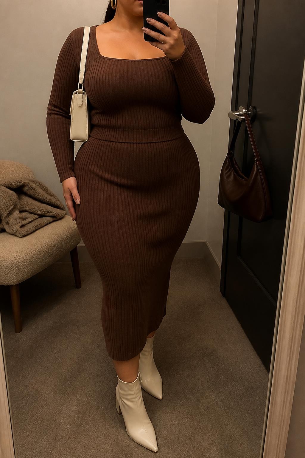 Mirror selfie of a plus size woman wearing a chocolate brown ribbed knit top with long sleeves and a square neckline, a matching chocolate brown high-waisted ribbed knit midi skirt, cream pointed ankle boots with low block heels, a cream structured shoulder bag, and chunky gold hoop earrings, clearly showing the exact matching knit set, brown ribbed texture, cream footwear, and accessories listed, holding a phone in front of a mirror, cropped at the neck so the face does not appear, relaxed elegant selfie pose, in a minimal dressing room with warm moody lighting, a handbag hanging on a door handle and a folded blanket on a chair, realistic modern Thanksgiving outfit, latest iPhone photo quality.