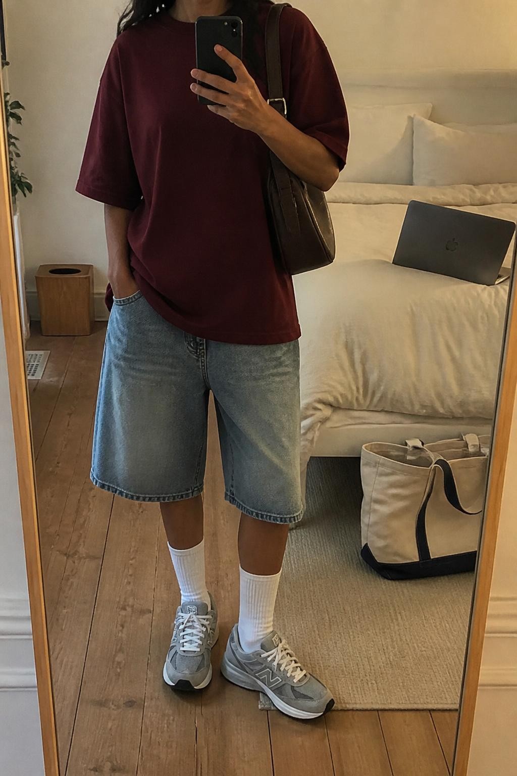 mirror selfie of a woman with warm tan skin wearing faded loose blue denim baggy jorts, an oversized burgundy heavyweight cotton T-shirt with no graphics, white crew socks, gray retro sneakers and a dark brown leather shoulder bag, wearing exactly the listed outfit with no added clothing or accessories, holding a phone in front of a mirror, cropped at NECK so the face does not show, relaxed casual selfie pose, in a cozy bedroom mirror setup, soft indoor lighting, laptop on the bed and a tote bag resting on the floor, natural slightly imperfect styling, latest iPhone photo quality.