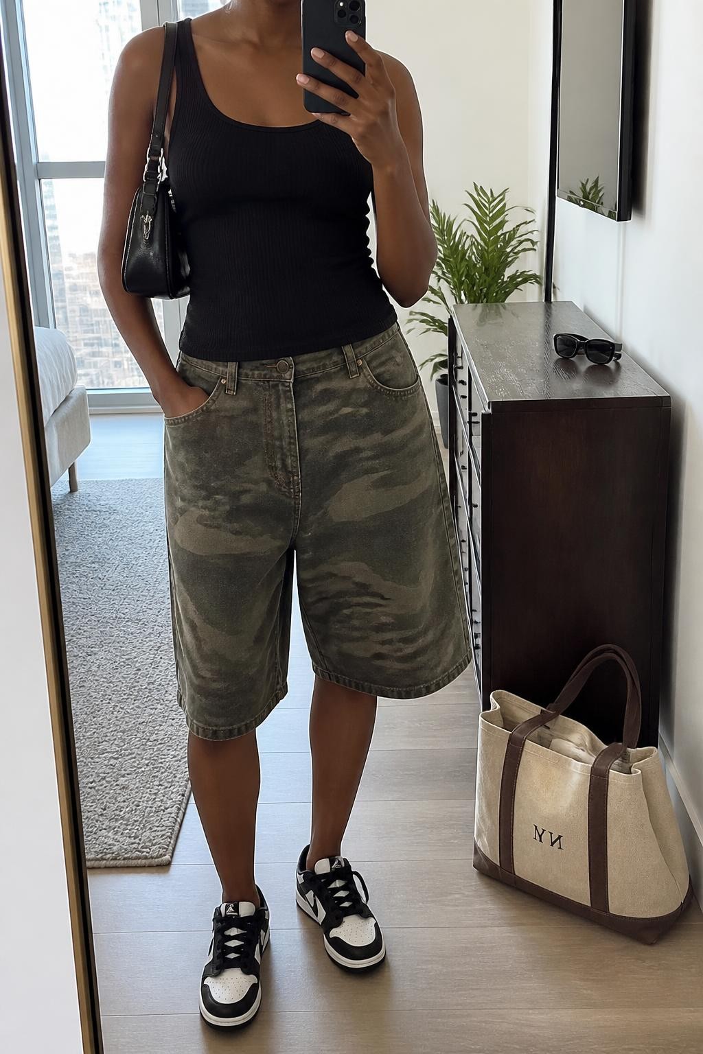 mirror selfie of a Black woman wearing loose knee-length green and brown camouflage denim jorts, a fitted black ribbed cotton tank top, black and white retro sneakers, a compact black nylon shoulder bag and medium silver hoop earrings, wearing exactly the listed clothing and accessories with no extra garments, holding a phone in front of a mirror, cropped at NECK so the face does not show, relaxed casual selfie pose, in a stylish city apartment, natural window light, sunglasses placed on a dresser and a tote bag resting on the floor, realistic denim texture and natural proportions, latest iPhone photo quality.