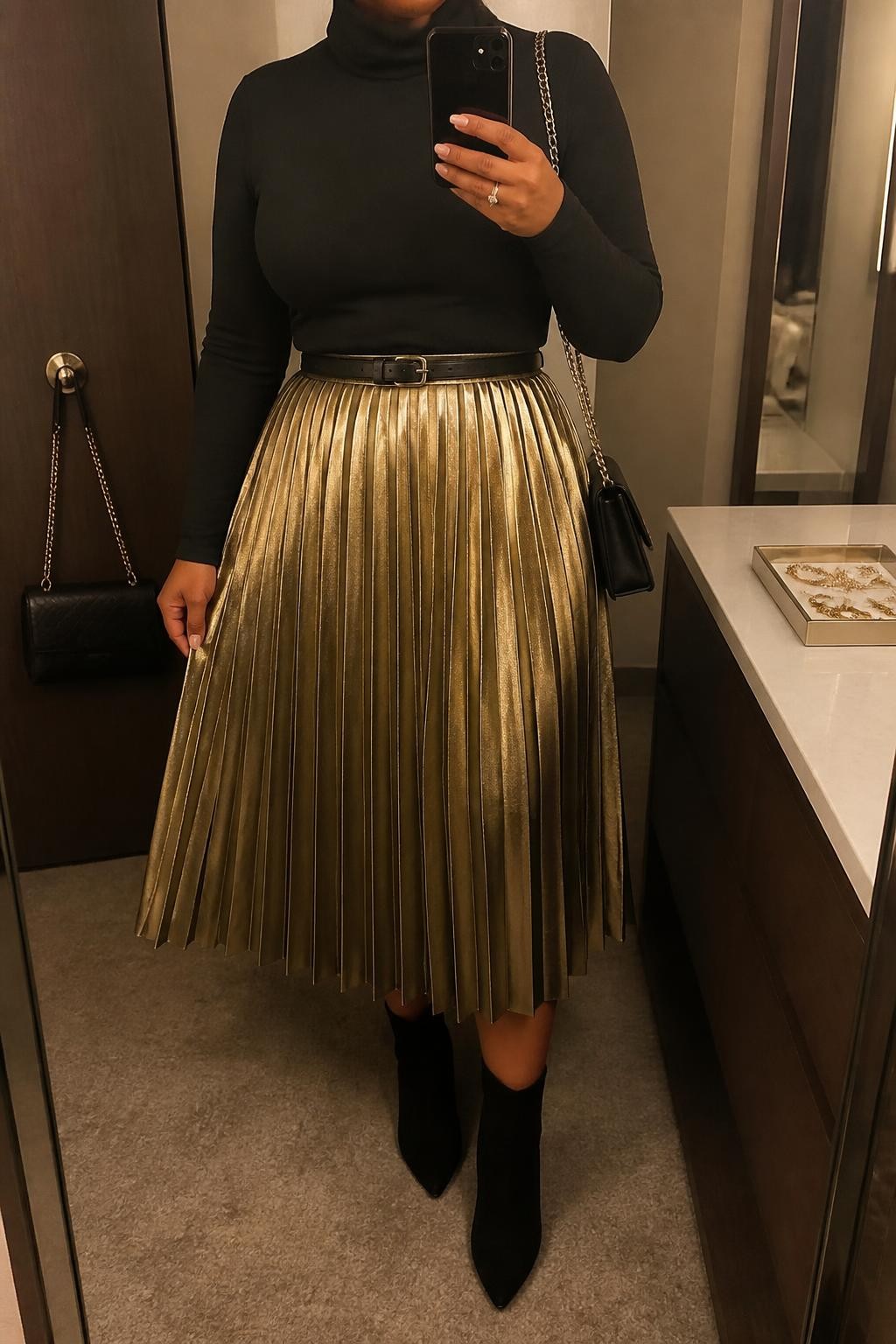 Mirror selfie of a plus size woman wearing a gold metallic pleated midi skirt, a fitted black long-sleeve turtleneck, a slim black leather belt, black suede ankle boots, and a small black chain-strap bag, clearly showing the exact clothing items, metallic texture, colors, fabrics, and accessories listed, holding a phone in front of a mirror, cropped at the neck so her face does not show, natural polished selfie pose, in a minimal dressing room with warm moody lighting, a handbag hanging on a door handle and a jewelry tray on the vanity, realistic elegant Christmas dinner appearance, latest iPhone photo quality.