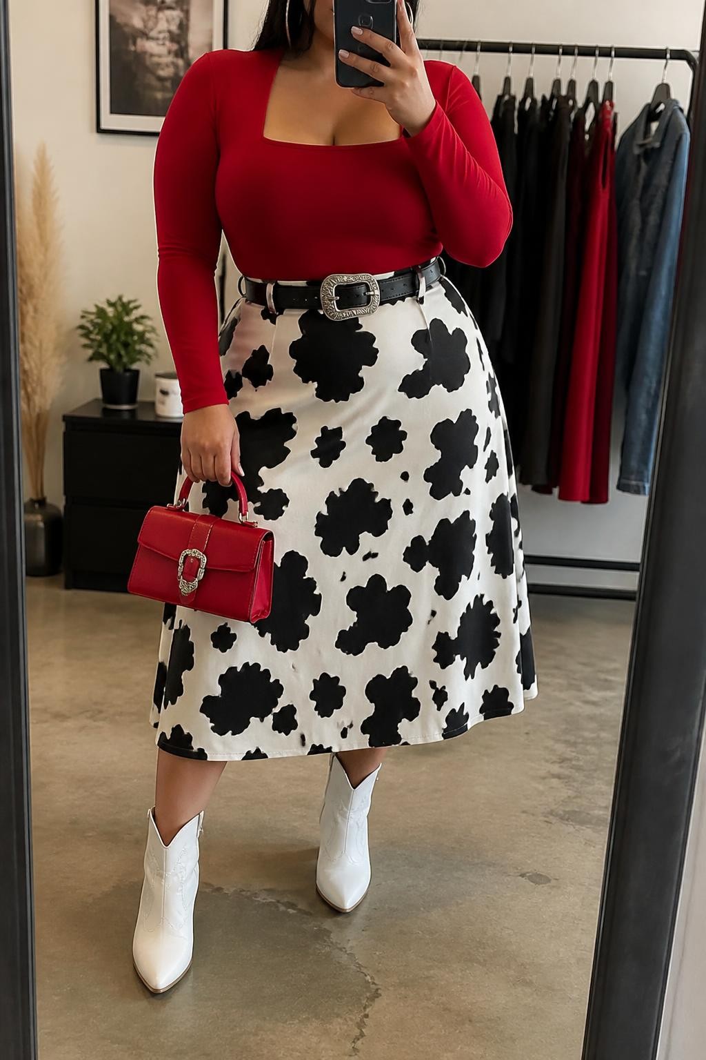 Mirror selfie of a plus size woman wearing a black and white cow-print high-waisted A-line midi skirt, a fitted red long-sleeve square-neck bodysuit, white pointed ankle cowboy boots, a black leather belt with a large silver buckle, and a structured red mini handbag, clearly showing the exact clothing items, cow print, red fitted top, white footwear, belt, and accessories listed, holding a phone in front of a mirror, cropped at the neck so the face does not show, relaxed fierce selfie stance, in a fashion studio mirror corner with soft indoor lighting, a clothing rack with garments behind her and a small plant on a dresser, realistic statement vaquera outfit, latest iPhone photo quality.