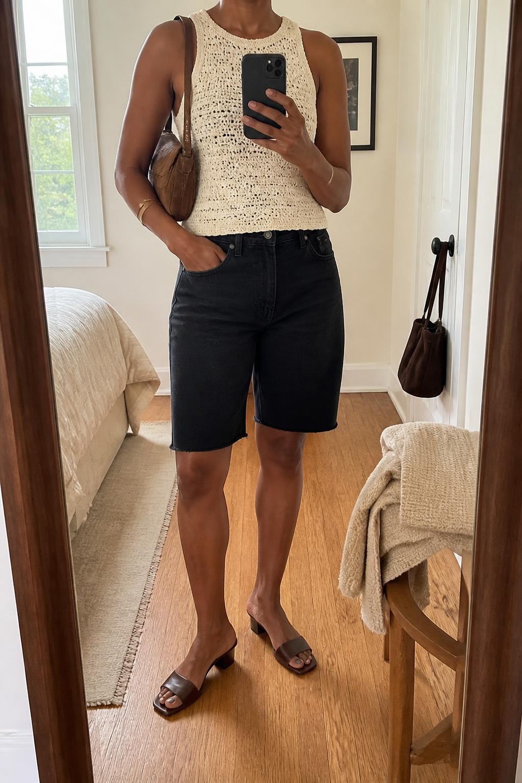 mirror selfie of a Black woman wearing clean black denim jorts with a straight knee-length fit, a fitted cream crochet sleeveless top, chocolate brown heeled mules with a low block heel, a brown suede shoulder bag and a simple gold cuff bracelet, wearing exactly the listed outfit with no additional garments or accessories, holding a phone in front of a mirror, cropped at NECK so the face does not show, relaxed casual selfie pose, in a cozy bedroom mirror setup, natural window light, folded blanket on a chair and a handbag hanging on a door handle, natural warm-weather styling, latest iPhone photo quality.