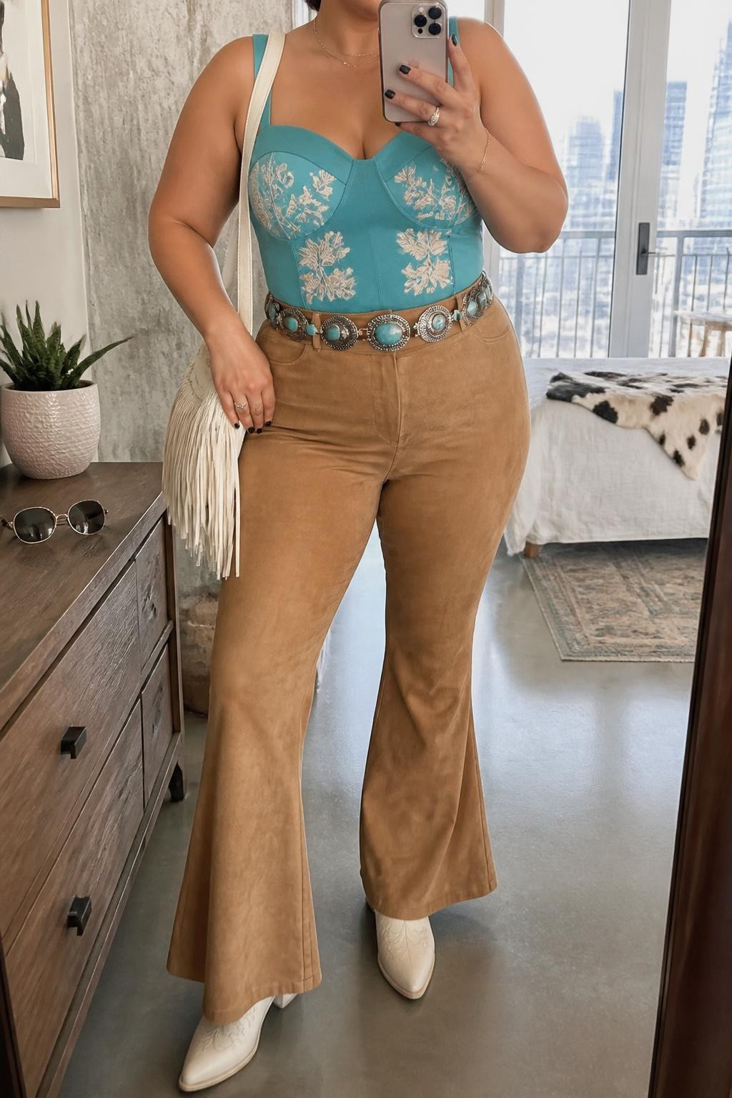 Mirror selfie of a plus size woman wearing tan suede high-waisted flared pants, a fitted turquoise camisole with wide supportive straps and cream floral embroidery, cream pointed cowboy boots, a turquoise concho belt with silver details, and a small cream fringe handbag, clearly showing the exact clothing items, tan suede, turquoise embroidery, cream boots, belt, and accessories listed, holding a phone in front of a mirror, cropped at the neck so the face does not appear, relaxed bold selfie stance, in a stylish city apartment with natural window light, sunglasses placed on a dresser and a small plant nearby, realistic warm-weather vaquera outfit, latest iPhone photo quality.