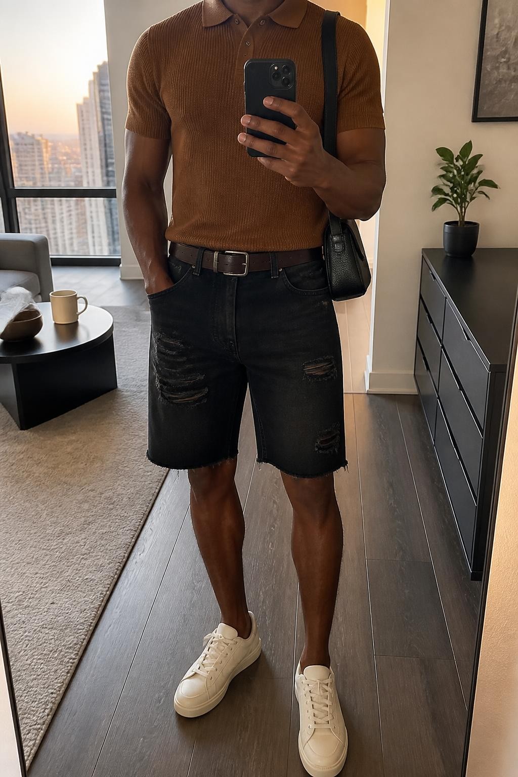 mirror selfie of a Black man wearing distressed black denim jorts with subtle frayed hems, a fitted rust brown short-sleeve knit polo, cream leather sneakers, a dark brown leather belt and a small black shoulder bag, wearing exactly the listed clothing and accessories with nothing additional, holding a phone in front of a mirror, cropped at NECK so the face does not show, relaxed casual selfie pose, in a stylish city apartment, evening golden hour, coffee mug on a side table and a small plant on a dresser, natural latest iPhone photo quality.