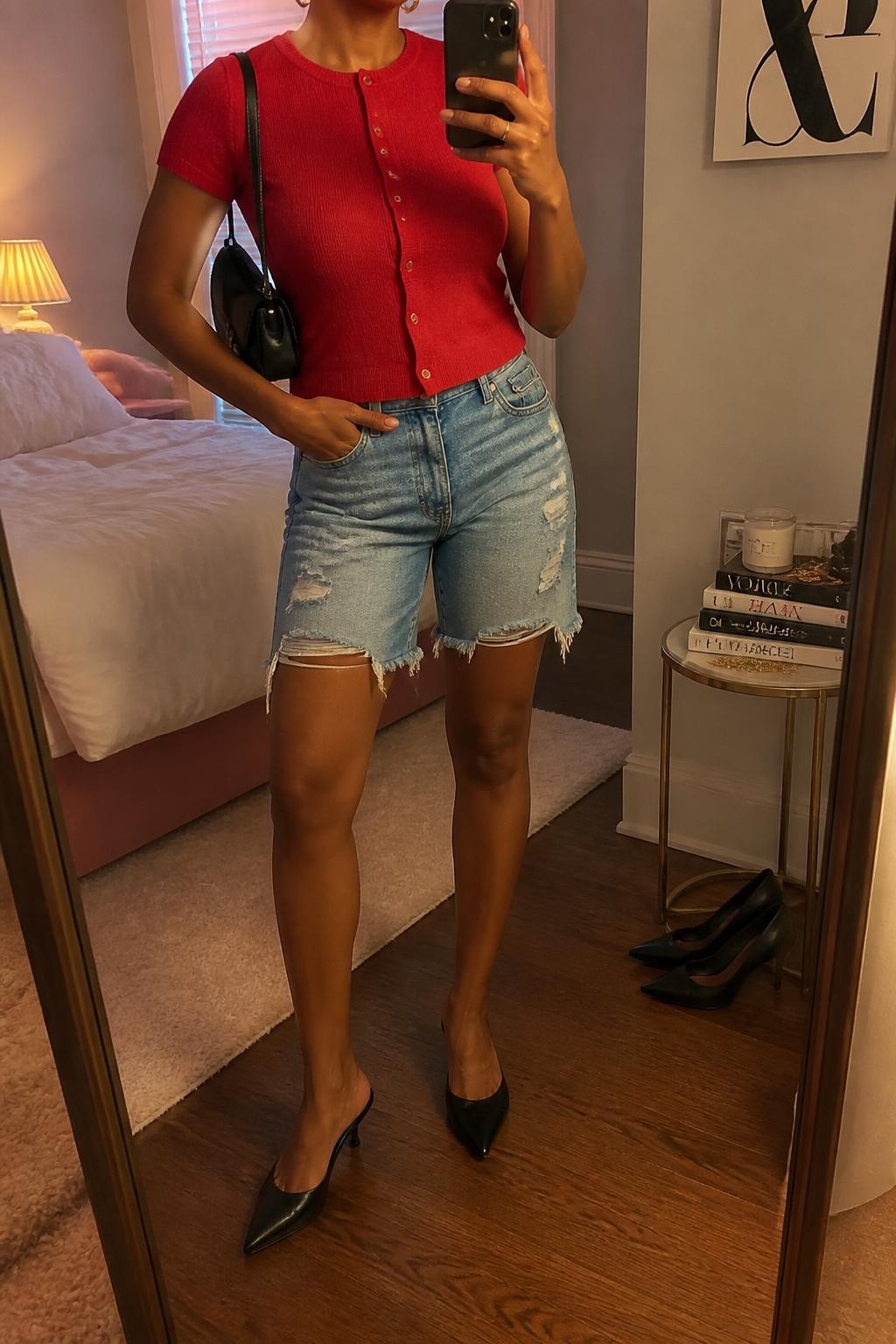 mirror selfie of a Black woman wearing distressed medium-blue denim jorts with lightly frayed hems, a fitted red fine-knit short-sleeve cardigan buttoned as a top, black pointed pumps with a low slim heel, a compact black leather shoulder bag and small gold hoop earrings, wearing exactly the listed clothing and accessories with no extra garments, holding a phone in front of a mirror, cropped at NECK so the face does not show, relaxed statement selfie pose, in a feminine-themed bedroom, warm moody lighting, pair of heels on the floor nearby and a stack of fashion magazines on a side table, believable latest iPhone photo quality.