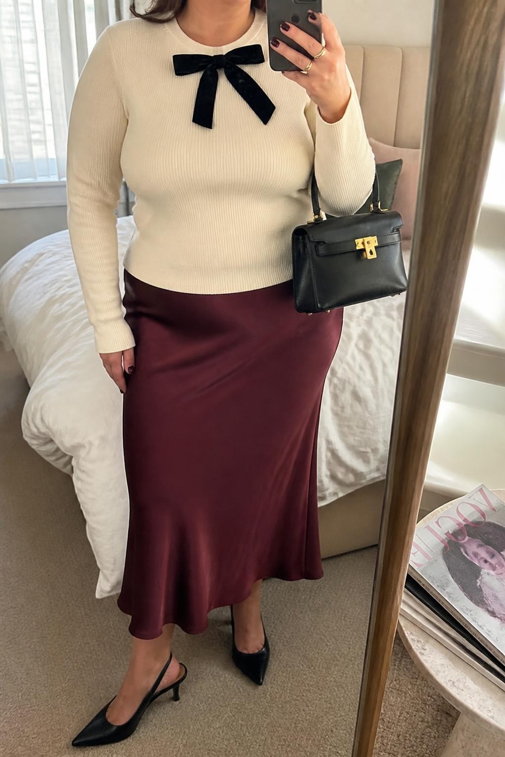 Mirror selfie of a plus size woman wearing a fluid burgundy satin midi skirt, a fitted cream knit sweater with a small black velvet bow at the neckline, black slingback heels, and a structured black mini bag, clearly showing the exact clothing items, colors, fabrics, bow detail, and accessories listed, holding a phone in front of a mirror, cropped at the neck so the face does not show, relaxed feminine selfie pose, in a cozy bedroom mirror setup with natural window light, soft pillows on an unmade bed and a stack of fashion magazines on a side table, realistic Christmas lunch outfit, latest iPhone photo quality.