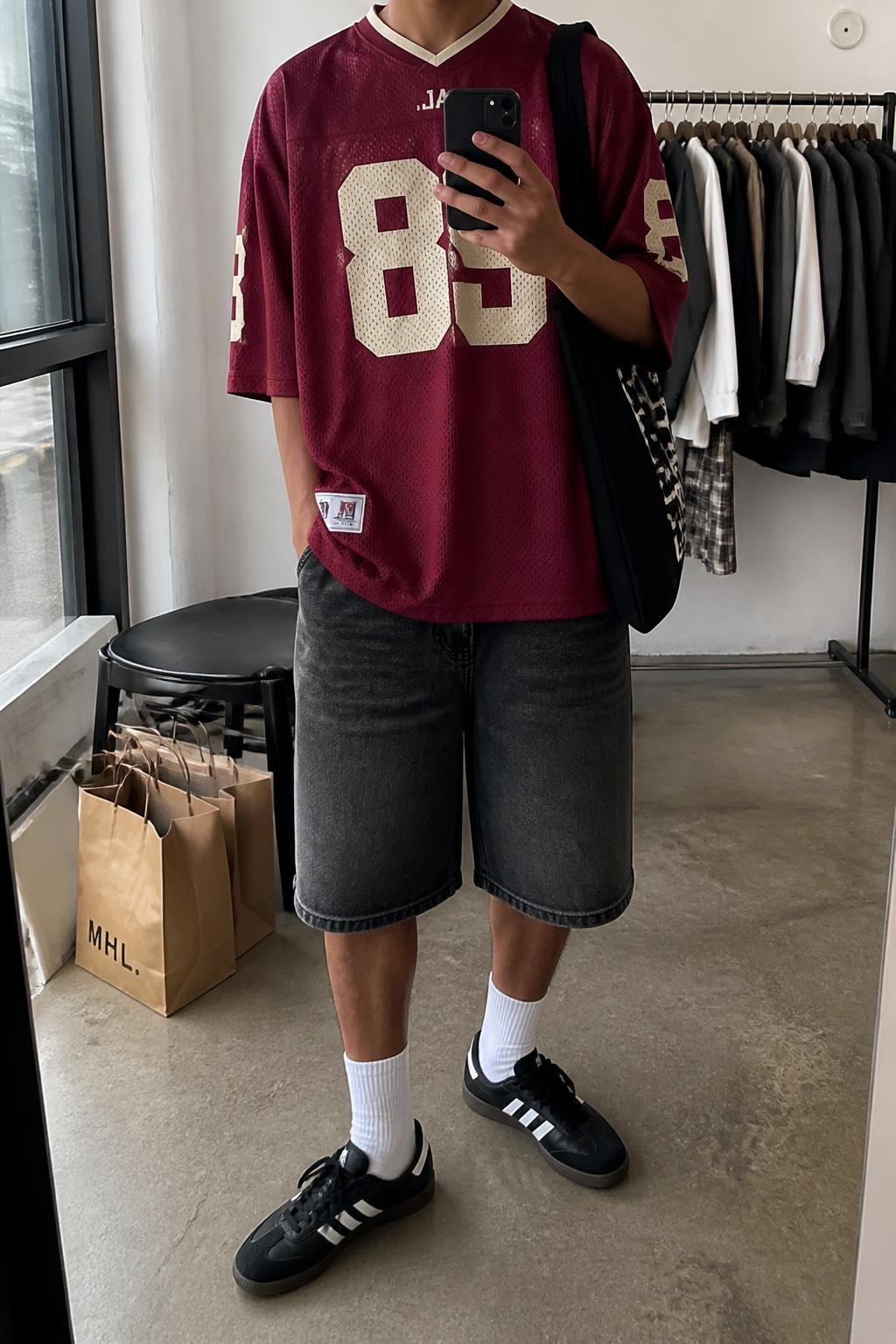 mirror selfie of a man with medium brown skin wearing faded black loose-fit denim jorts, an oversized burgundy football jersey with simple cream number detailing, white crew socks, black retro sneakers and a black canvas tote bag, wearing exactly the listed outfit with no added garments, holding a phone in front of a mirror, cropped at NECK so the face does not show, relaxed masculine selfie pose, in a fashion studio mirror corner, natural window light, clothing rack with garments behind and shopping bags beside a chair, realistic casual streetwear appearance, latest iPhone photo quality.