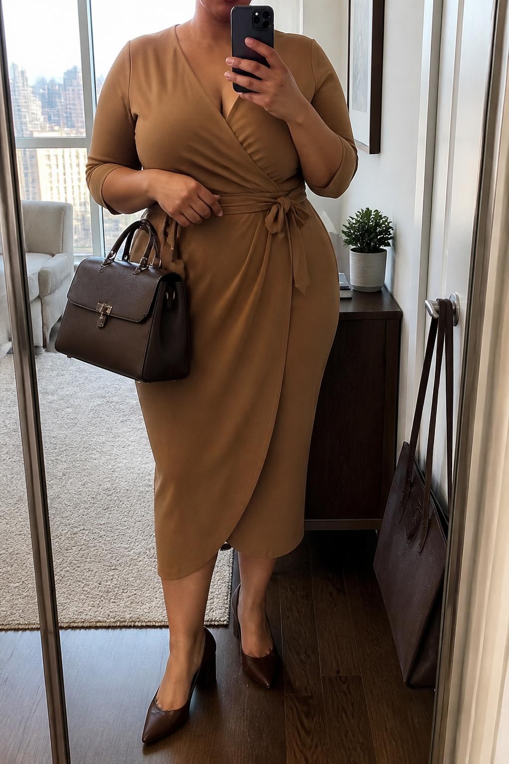 Mirror selfie of a plus size woman wearing a camel brown crepe wrap midi dress with three-quarter sleeves, a modest V-neckline, and a matching waist tie, chocolate brown pointed block-heel pumps, a structured chocolate leather handbag, and small gold stud earrings, clearly showing the exact clothing items, camel dress, wrap shape, colors, footwear, and accessories listed, holding a phone in front of a mirror, cropped at the neck so the face does not show, relaxed professional selfie pose, in a stylish city apartment with natural window light, a handbag hanging on a door handle and a small plant on a dresser, realistic interview appearance, latest iPhone photo quality.