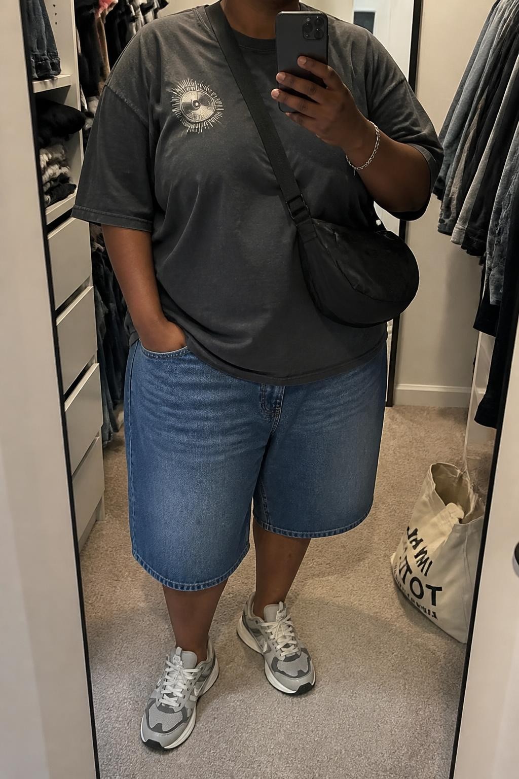 mirror selfie of a plus size Black woman wearing baggy blue denim jorts with a loose knee-length cut, an oversized charcoal gray cotton graphic T-shirt with a small faded cream graphic, gray and white retro sneakers, a black nylon crossbody bag and a silver chain bracelet, wearing exactly the listed outfit with no additional clothing or accessories, holding a phone in front of a mirror, cropped at NECK so the face does not show, relaxed street-style selfie pose, in a walk-in closet mirror, soft indoor lighting, open closet with clothes visible and a tote bag resting on the floor, natural slightly relaxed styling, latest iPhone photo quality.