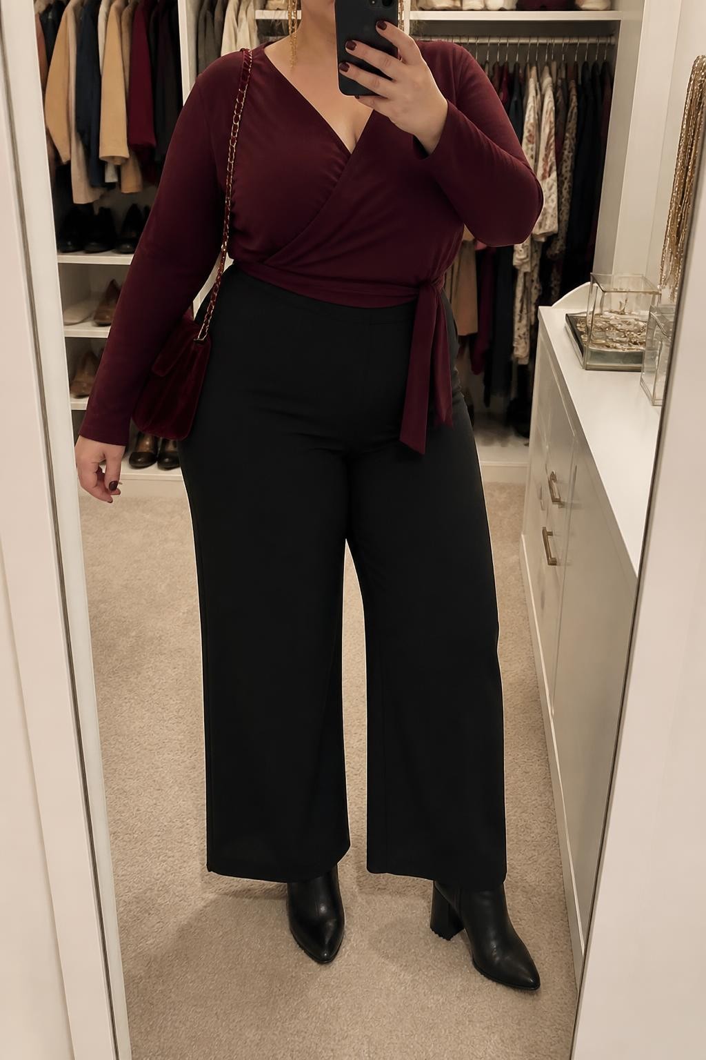 Mirror selfie of a plus size woman wearing a burgundy long-sleeve jersey wrap top with a softly tied waist, high-waisted black wide-leg trousers in stretch crepe, black pointed block-heel ankle boots, a burgundy velvet shoulder bag, and delicate gold chain earrings, clearly showing the exact clothing items, burgundy jersey, black trousers, footwear, and accessories listed, holding a phone in front of a mirror, cropped at the neck so the face is hidden, relaxed polished selfie pose, in a walk-in closet mirror with soft indoor lighting, an open closet with clothes visible and a jewelry tray on the vanity, realistic dressy Thanksgiving appearance, latest iPhone photo quality.
