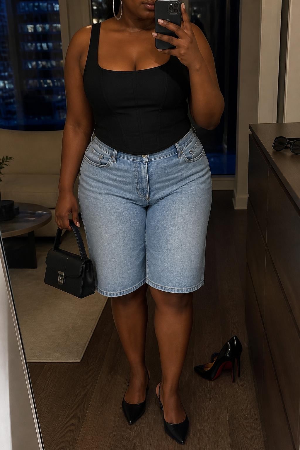 mirror selfie of a plus size Black woman wearing light blue relaxed denim jorts with a knee-length fit, a fitted black cotton corset-inspired top with clean seam detailing, black pointed slingback flats, a structured black mini handbag and medium silver hoop earrings, wearing exactly the listed clothing and accessories with nothing additional, holding a phone in front of a mirror, cropped at NECK so the face does not show, relaxed confident selfie pose, in a stylish city apartment, warm moody lighting, pair of heels on the floor nearby and sunglasses placed on a dresser, realistic polished casual styling, latest iPhone photo quality.