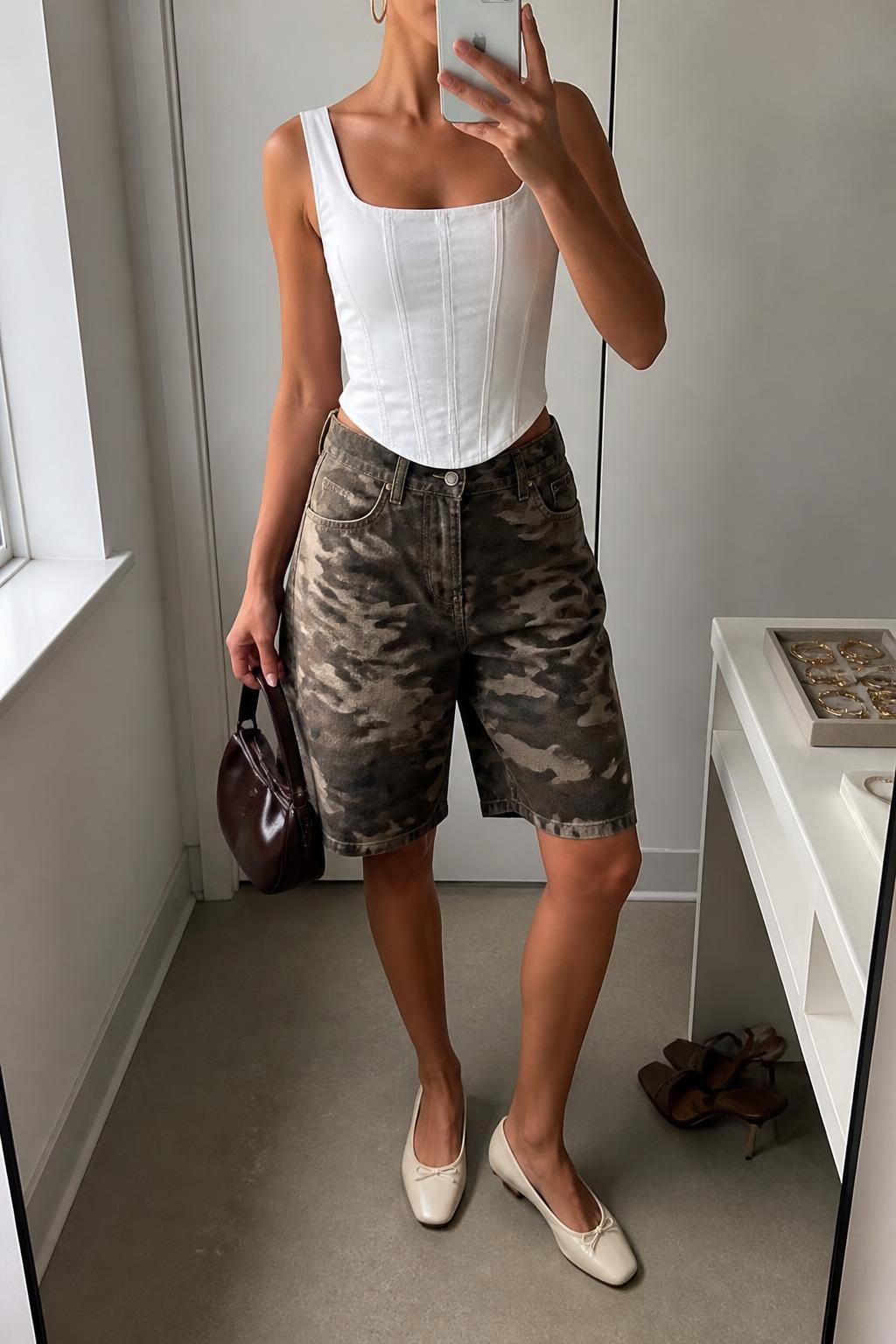 mirror selfie of a woman with warm tan skin wearing brown and olive camouflage denim jorts with a relaxed knee-length fit, a fitted white cotton corset-inspired top with clean seam detailing, cream leather flats, a dark brown mini handbag and medium gold hoop earrings, wearing exactly the listed clothing and accessories with nothing additional, holding a phone in front of a mirror, cropped at NECK so the face does not show, relaxed selfie pose, in a minimal dressing room, natural window light, jewelry tray on the vanity and a pair of heels on the floor nearby, believable polished casual styling, latest iPhone photo quality.