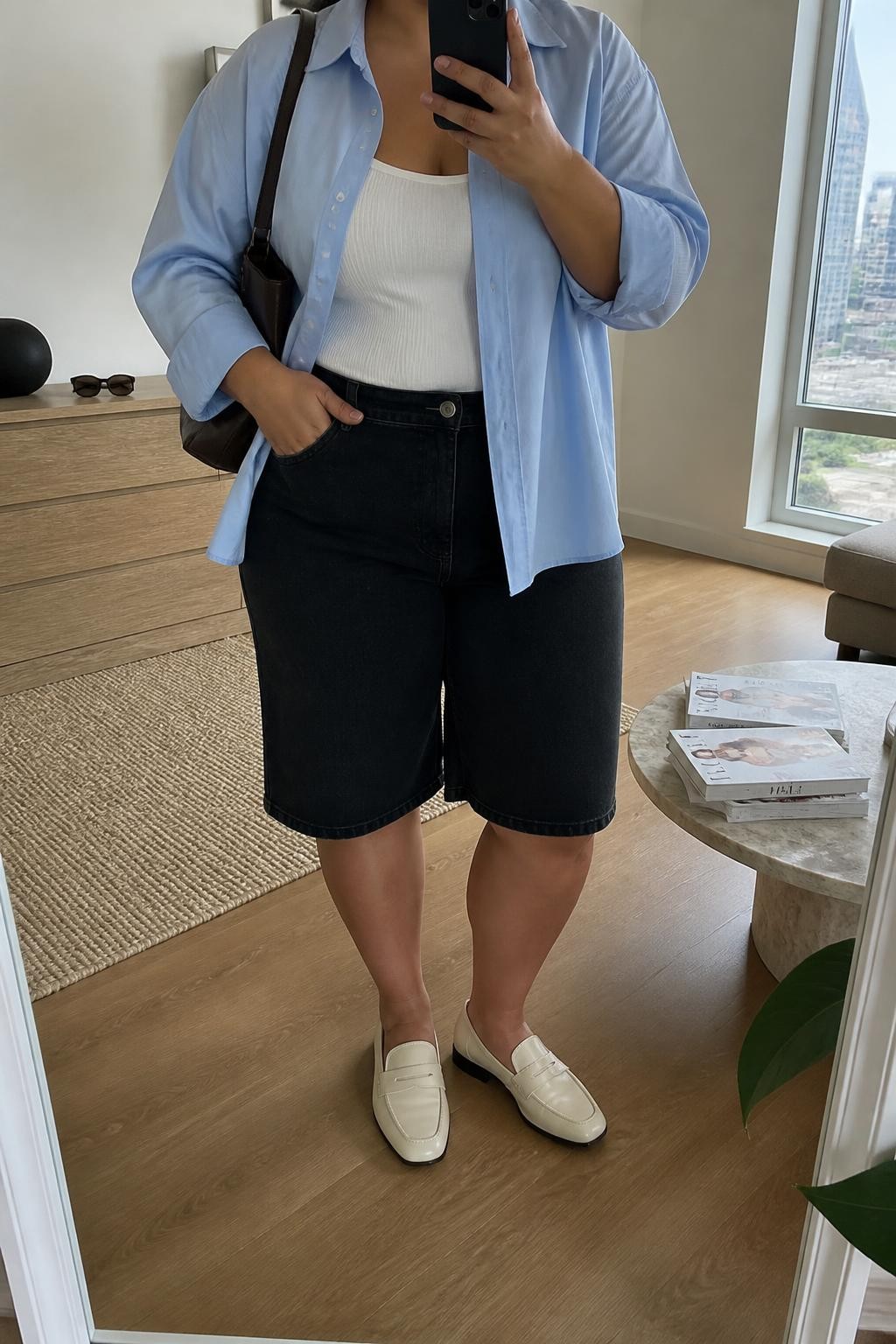 mirror selfie of a plus size woman with warm tan skin wearing long black denim jorts ending near the knee, a pale blue oversized cotton button-up shirt worn open over a fitted white ribbed tank, cream loafers and a dark brown leather shoulder bag, wearing exactly the listed outfit with no additional clothing pieces, holding a phone in front of a mirror, cropped at NECK so the face does not show, relaxed casual selfie pose, in a stylish city apartment, natural window light, sunglasses placed on a dresser and a stack of fashion magazines on a side table, realistic latest iPhone photo quality.