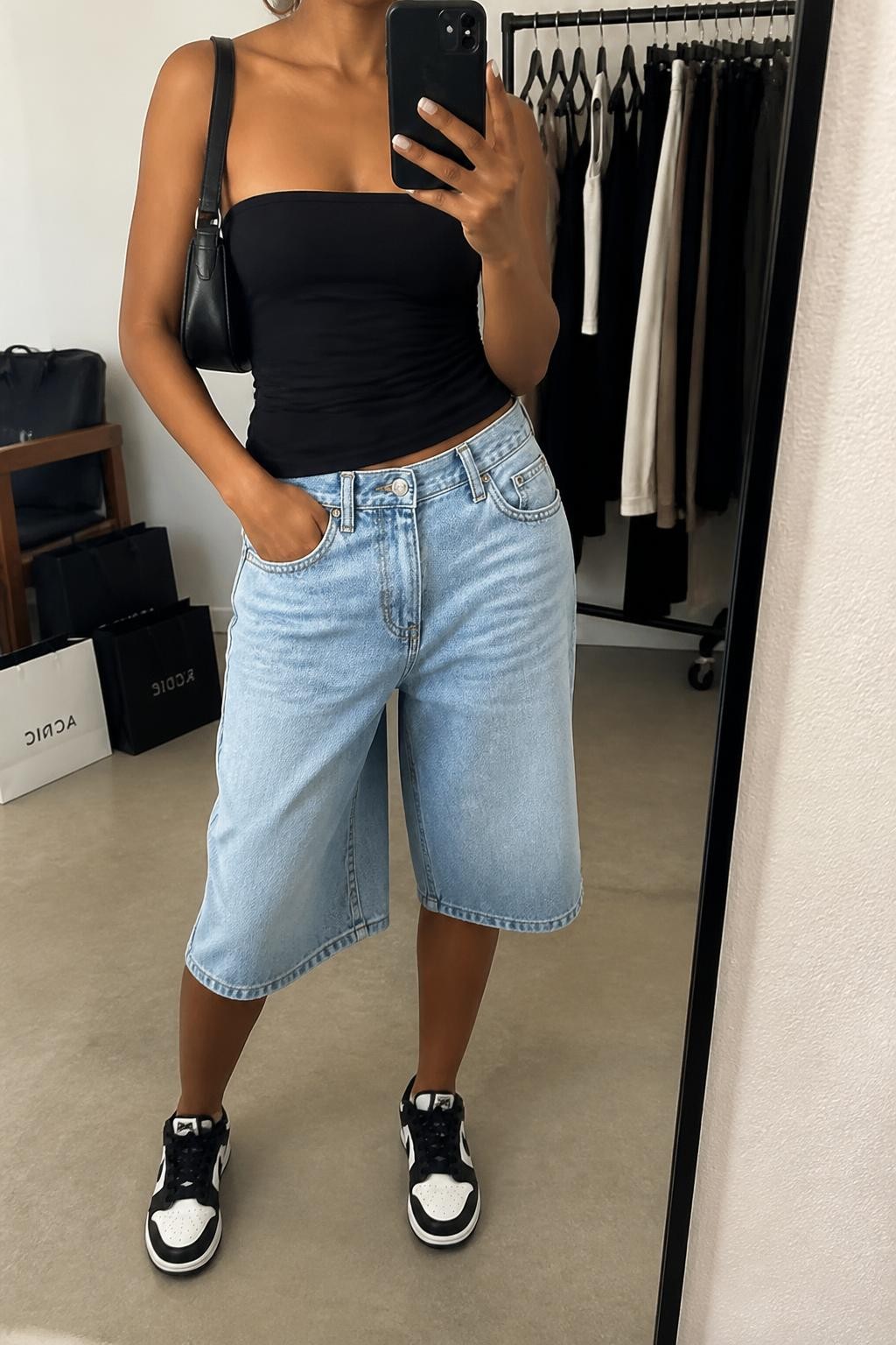 mirror selfie of a woman with medium brown skin wearing light-wash long denim jorts with a loose below-the-knee cut, a fitted black stretch cotton tube top, black and white retro sneakers, a black leather mini shoulder bag and chunky silver hoop earrings, wearing exactly the listed outfit with no additional clothing pieces, holding a phone in front of a mirror, cropped at NECK so the face does not show, relaxed casual selfie pose, in a fashion studio mirror corner, soft indoor lighting, clothing rack with garments behind and shopping bags beside a chair, natural cool-girl styling, latest iPhone photo quality.