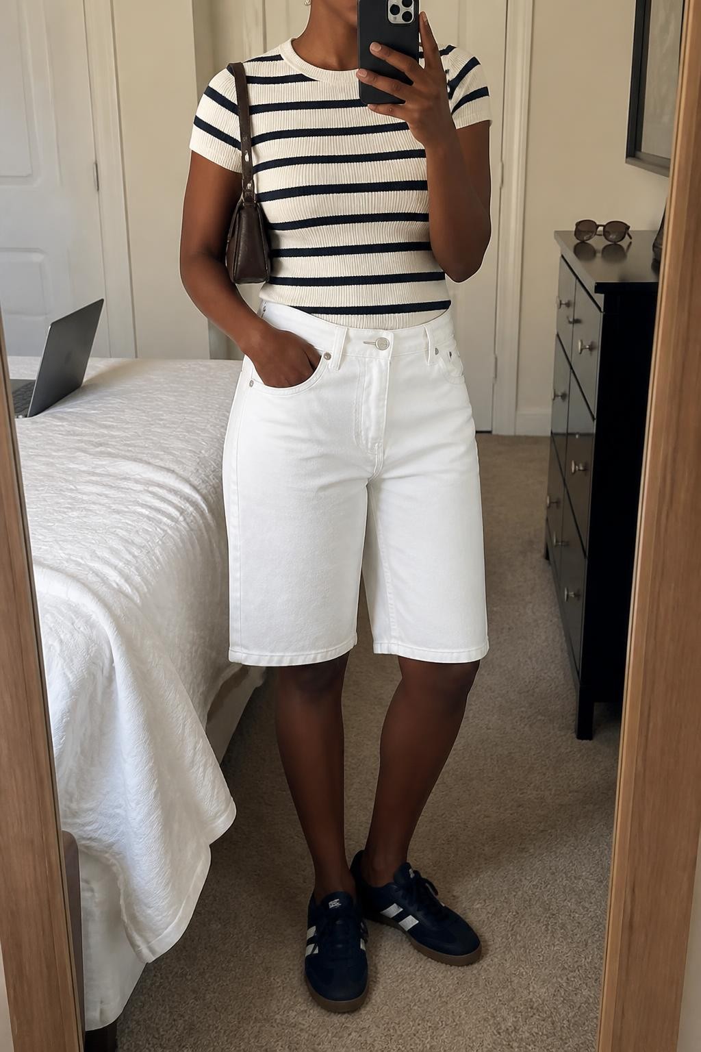 mirror selfie of a Black woman wearing clean white denim jorts with a relaxed knee-length fit, a fitted cream and navy striped short-sleeve knit top, navy low-profile sneakers, a dark brown mini shoulder bag and small gold stud earrings, wearing exactly the listed outfit with no added clothing pieces, holding a phone in front of a mirror, cropped at NECK so the face does not show, relaxed casual selfie pose, in a cozy bedroom mirror setup, soft indoor lighting, laptop on the bed and sunglasses placed on a dresser, natural latest iPhone photo quality.