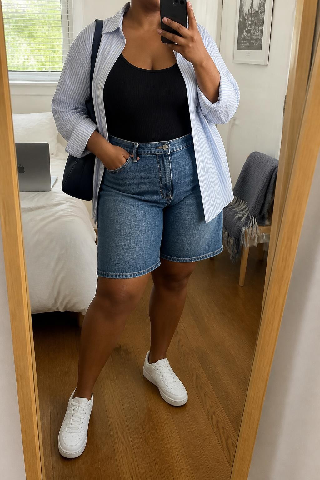 mirror selfie of a plus size Black woman wearing medium-wash straight-cut denim jorts, an oversized pale blue and white striped cotton shirt worn open over a fitted black ribbed tank, white leather sneakers, a navy shoulder bag and small silver hoop earrings, wearing exactly the listed outfit with no added garments, holding a phone in front of a mirror, cropped at NECK so the face does not show, relaxed casual selfie pose, in a cozy bedroom mirror setup, natural window light, laptop on the bed and a folded blanket on a chair, believable everyday styling, latest iPhone photo quality.