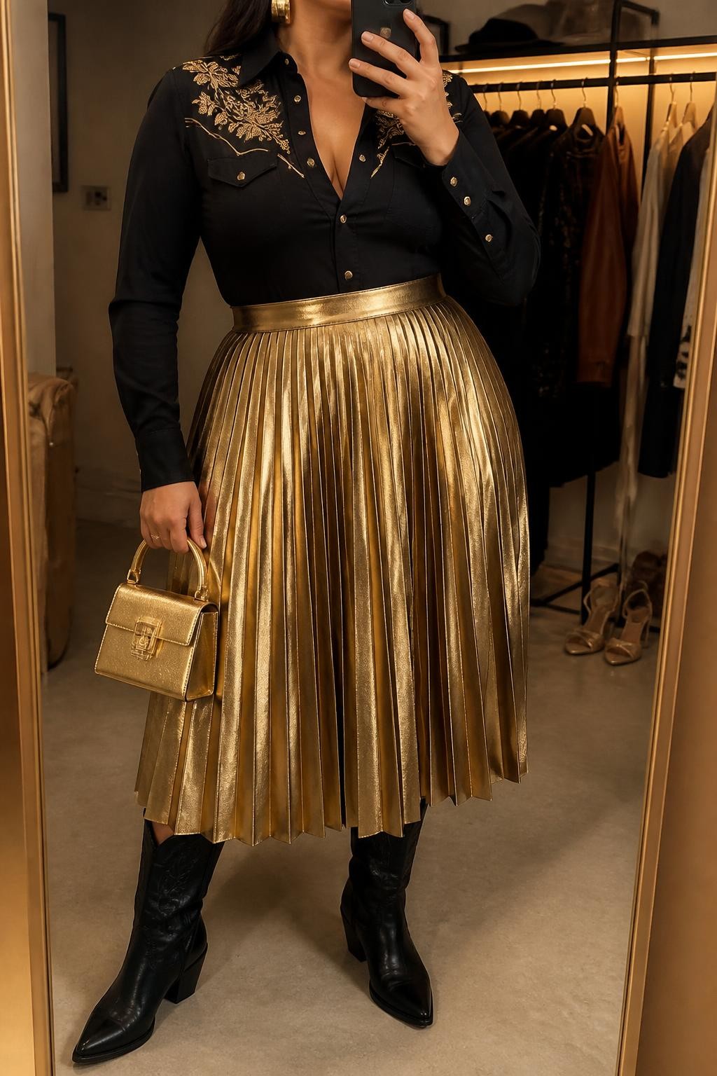Mirror selfie of a plus size woman wearing a metallic gold high-waisted pleated midi skirt, a fitted black Western blouse with long sleeves, gold floral embroidery across the shoulders, and gold snap buttons, black leather knee-high cowboy boots, a structured gold mini handbag, and chunky gold cuff earrings, clearly showing the exact clothing items, metallic skirt, black blouse, gold embroidery, boots, bag, and accessories listed, holding a phone in front of a mirror, cropped at the neck so the face does not show, relaxed fierce selfie stance, in a fashion studio mirror corner with warm moody lighting, a clothing rack with garments behind her and a pair of heels on the floor nearby, realistic show-stopping vaquera outfit, latest iPhone photo quality.
