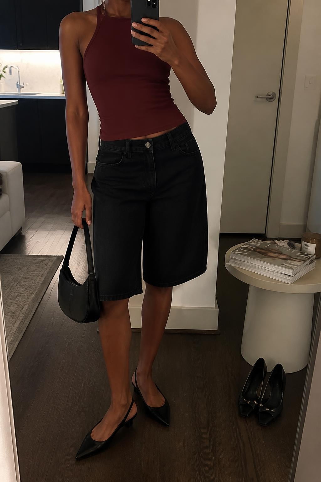 mirror selfie of a Black woman wearing clean long black denim jorts with a straight below-the-knee fit, a fitted burgundy halter top in stretch fabric, pointed black slingback flats, a small black leather handbag and slim silver drop earrings, wearing exactly the listed outfit with no additional garments, holding a phone in front of a mirror, cropped at NECK so the face does not show, relaxed selfie pose, in a stylish city apartment, warm moody lighting, pair of heels on the floor nearby and a stack of fashion magazines on a side table, natural realistic latest iPhone photo quality.
