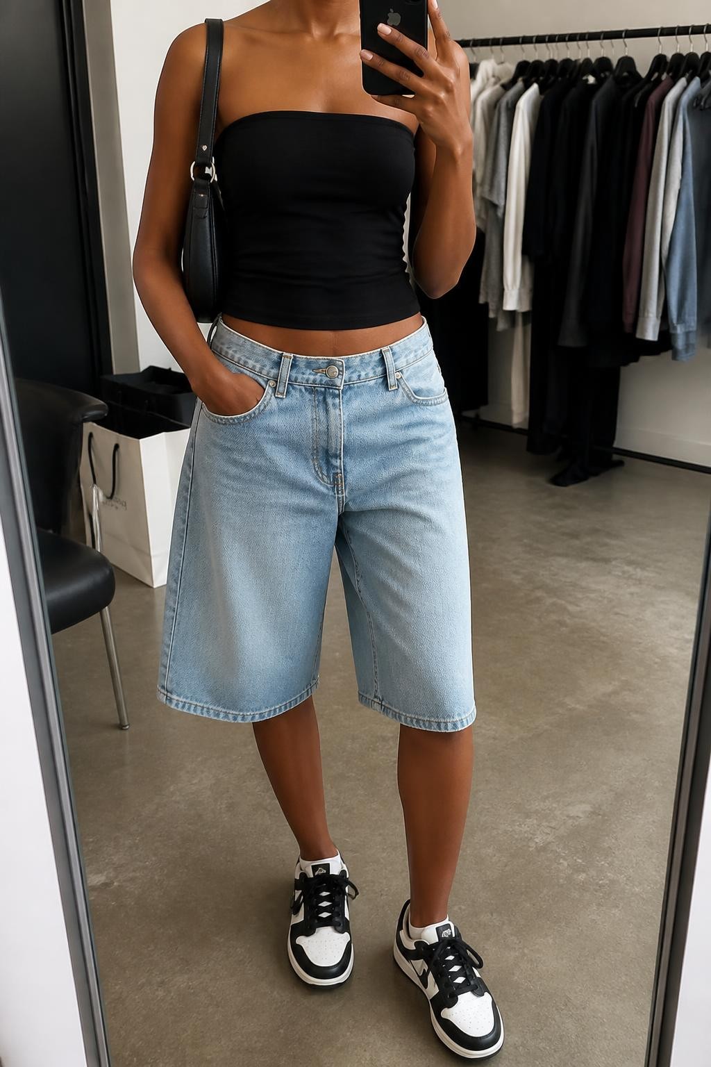 mirror selfie of a Black woman wearing loose light-wash denim baggy jorts ending just below the knee, a fitted black stretch cotton tube top, black and white retro sneakers, a black nylon shoulder bag and chunky silver hoop earrings, wearing exactly the listed clothing and accessories with nothing additional, holding a phone in front of a mirror, cropped at NECK so the face does not show, relaxed cool-girl selfie pose, in a fashion studio mirror corner, soft indoor lighting, clothing rack with garments behind and shopping bags beside a chair, natural fabric texture and latest iPhone photo quality.