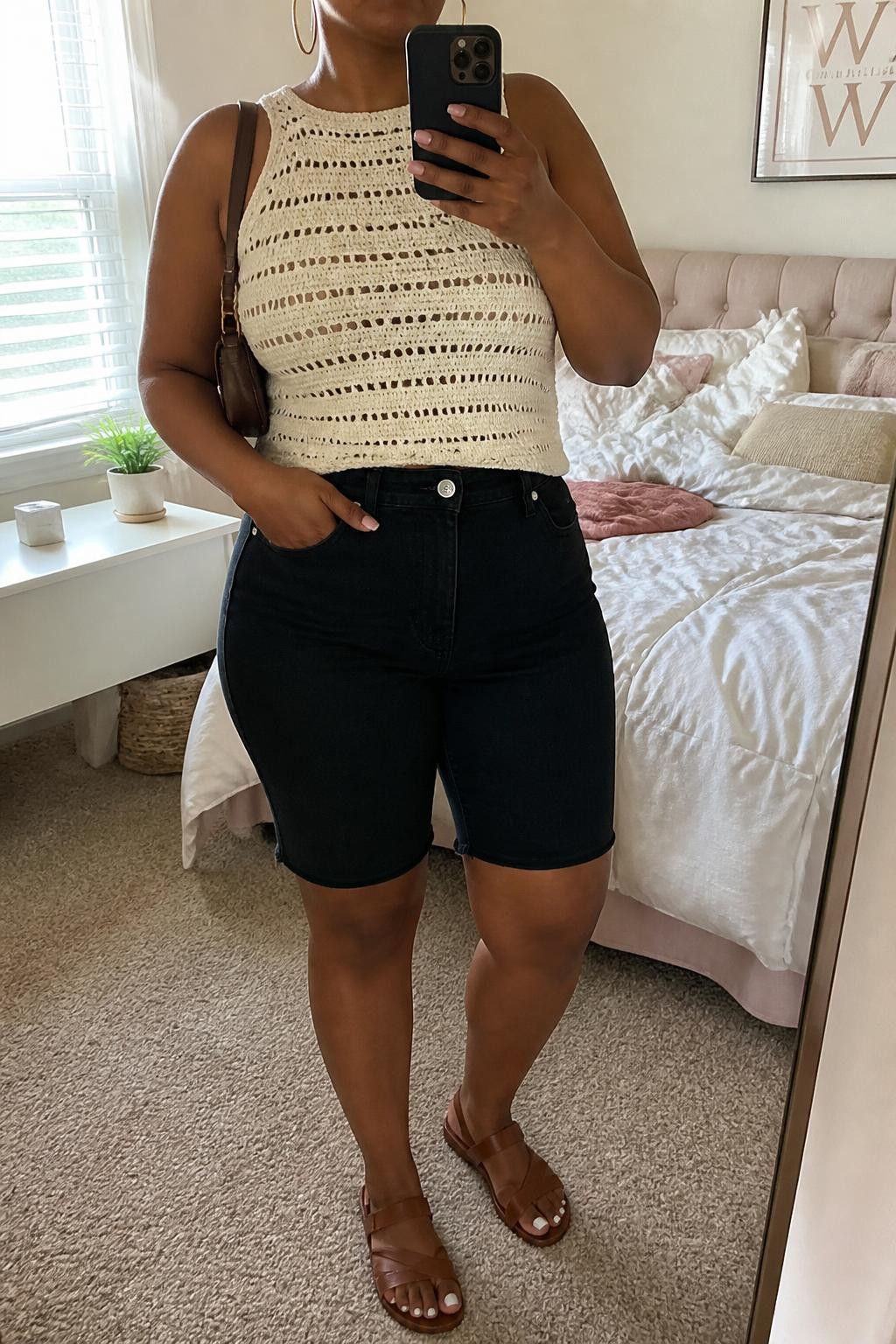 mirror selfie of a plus size Black woman wearing high-rise clean black denim jorts with a straight fit, a fitted sleeveless cream crochet top, brown leather sandals with flat straps, a brown mini shoulder bag and medium gold hoop earrings, wearing exactly the listed clothing and accessories without added garments, holding a phone in front of a mirror, cropped at NECK so the face does not show, relaxed casual selfie pose, in a feminine-themed bedroom, natural window light, soft pillows on an unmade bed and a small plant on a dresser, realistic warm-weather styling, latest iPhone photo quality.