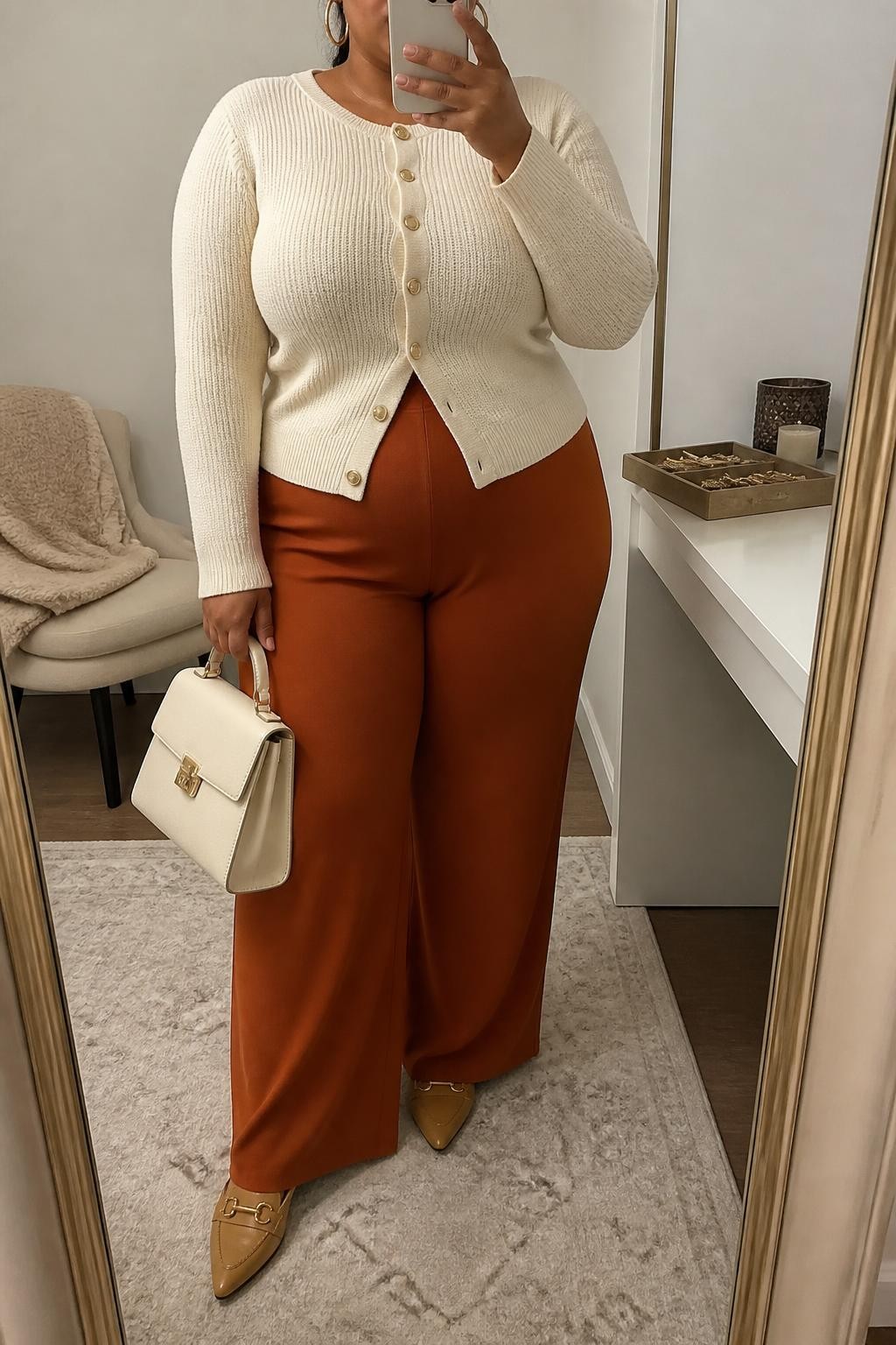 Mirror selfie of a plus size woman wearing a fitted cream knit cardigan buttoned at the front, rust orange high-waisted palazzo trousers in soft stretch fabric, tan pointed loafers, a structured cream handbag, and hammered gold hoop earrings, clearly showing the exact clothing items, cream knit, rust trousers, tan footwear, and accessories listed, holding a phone in front of a mirror, cropped at the neck so the face does not show, relaxed elegant selfie pose, in a fashion studio mirror corner with soft indoor lighting, a jewelry tray on the vanity and a folded blanket on a chair, realistic comfortable Thanksgiving outfit, latest iPhone photo quality.